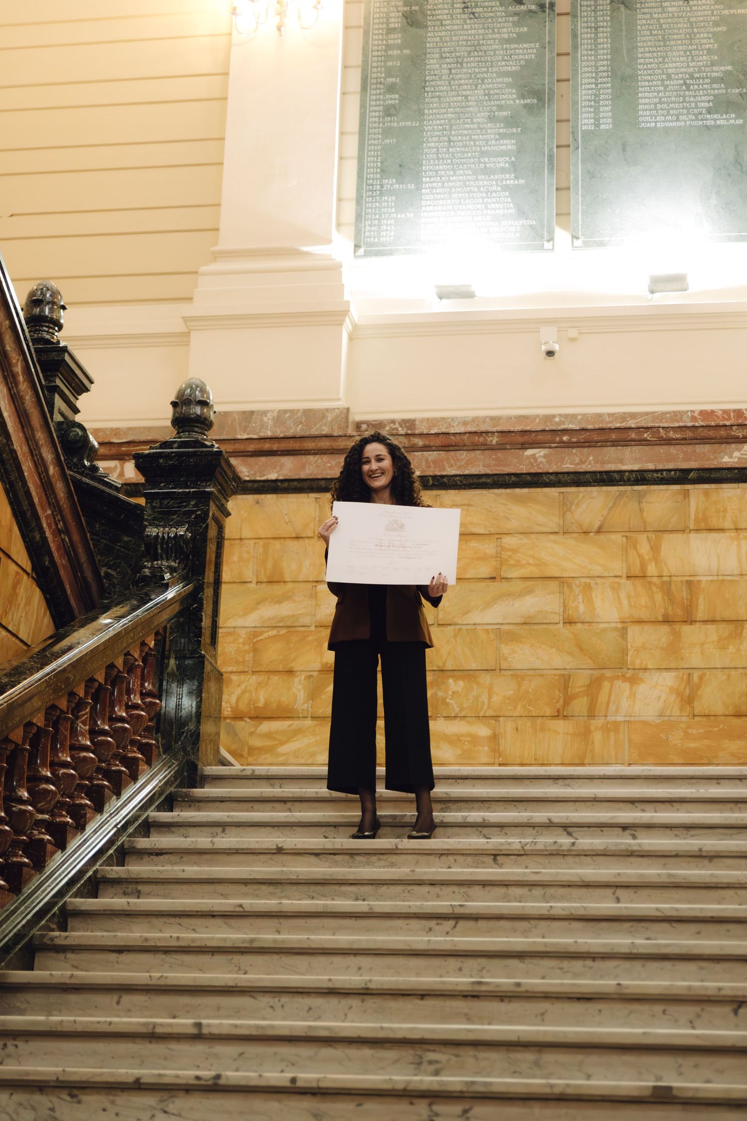Woman holding a certificate while standing on grand marble staircase in a historic building.