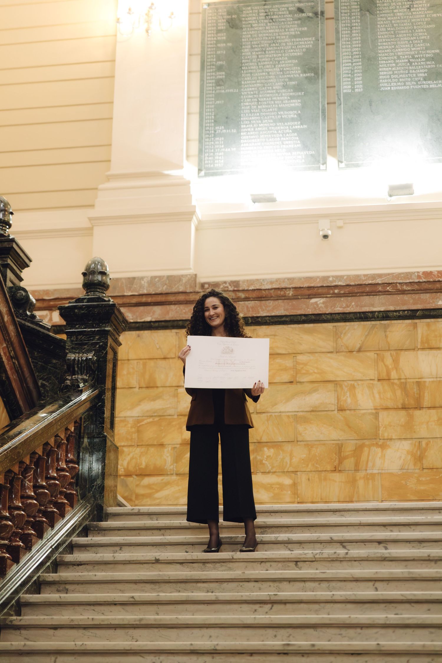 Smiling woman in brown blazer holding a certificate on marble stairs in a formal building.