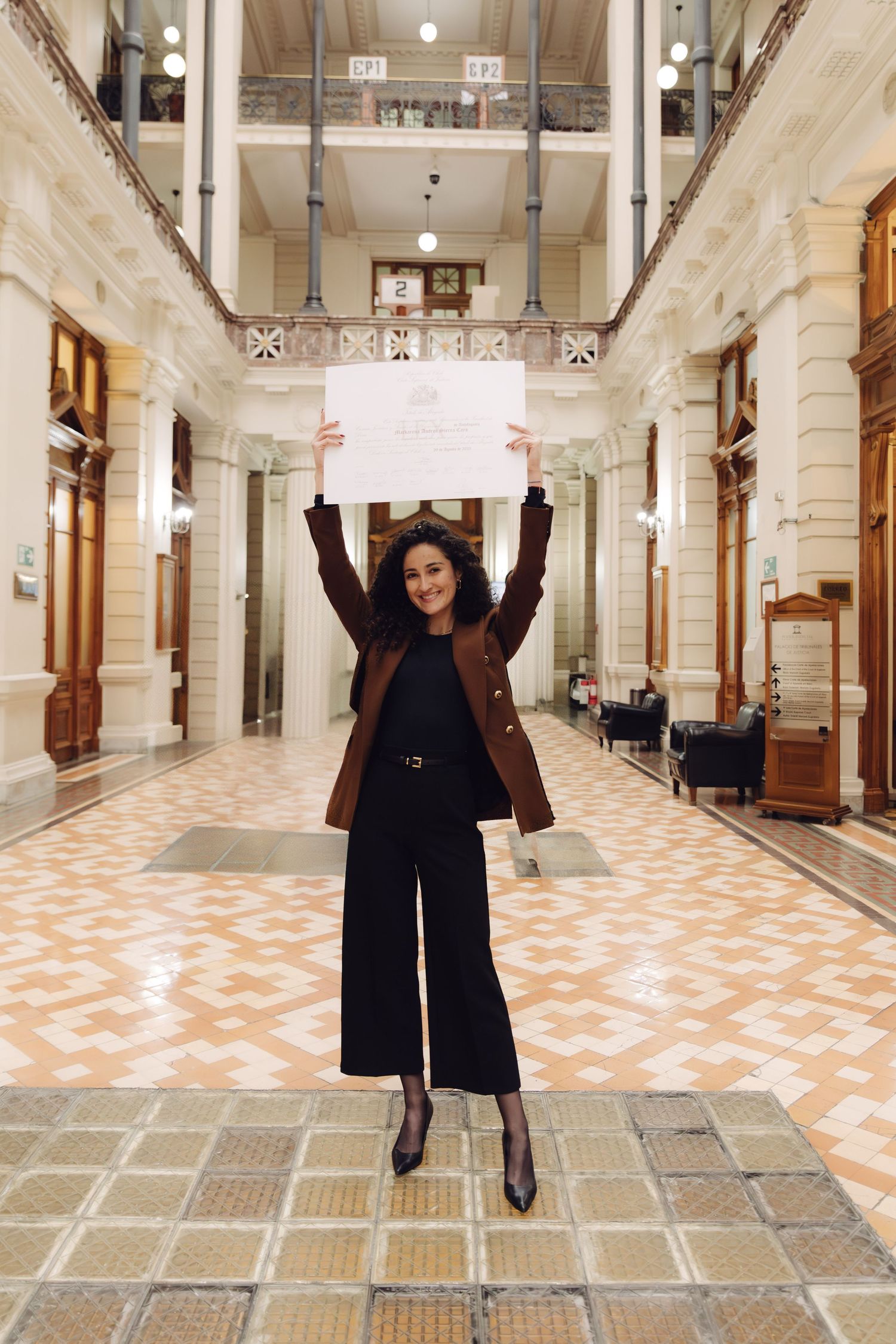 Joyful woman raising a certificate overhead in a grand historic building with tiled floors.