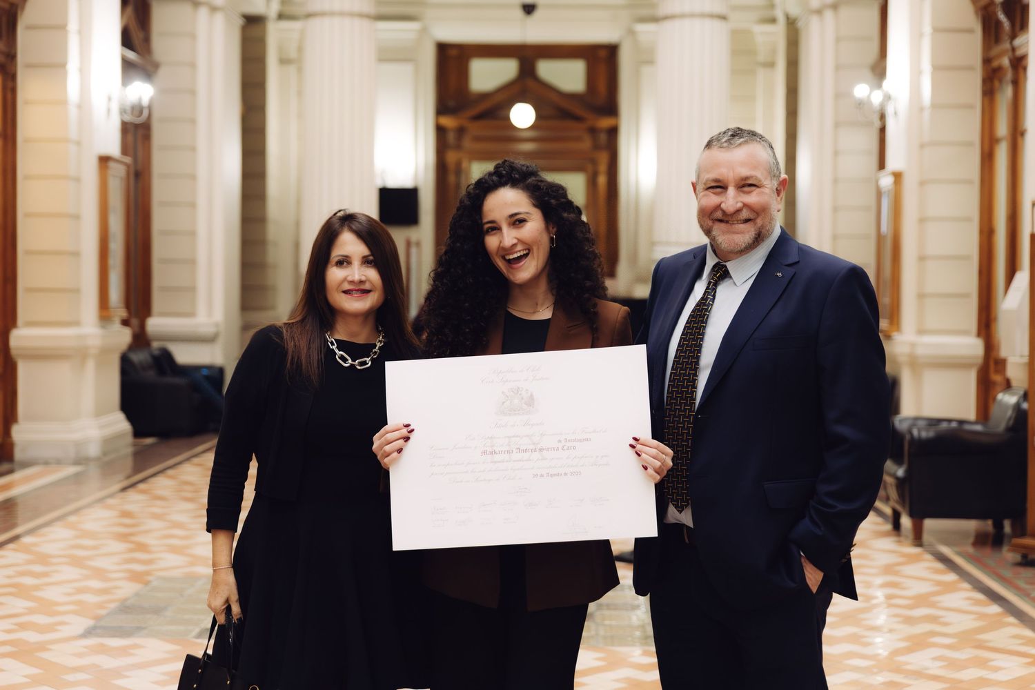 Three people posing with an official certificate in an elegant hall with ornate architecture.