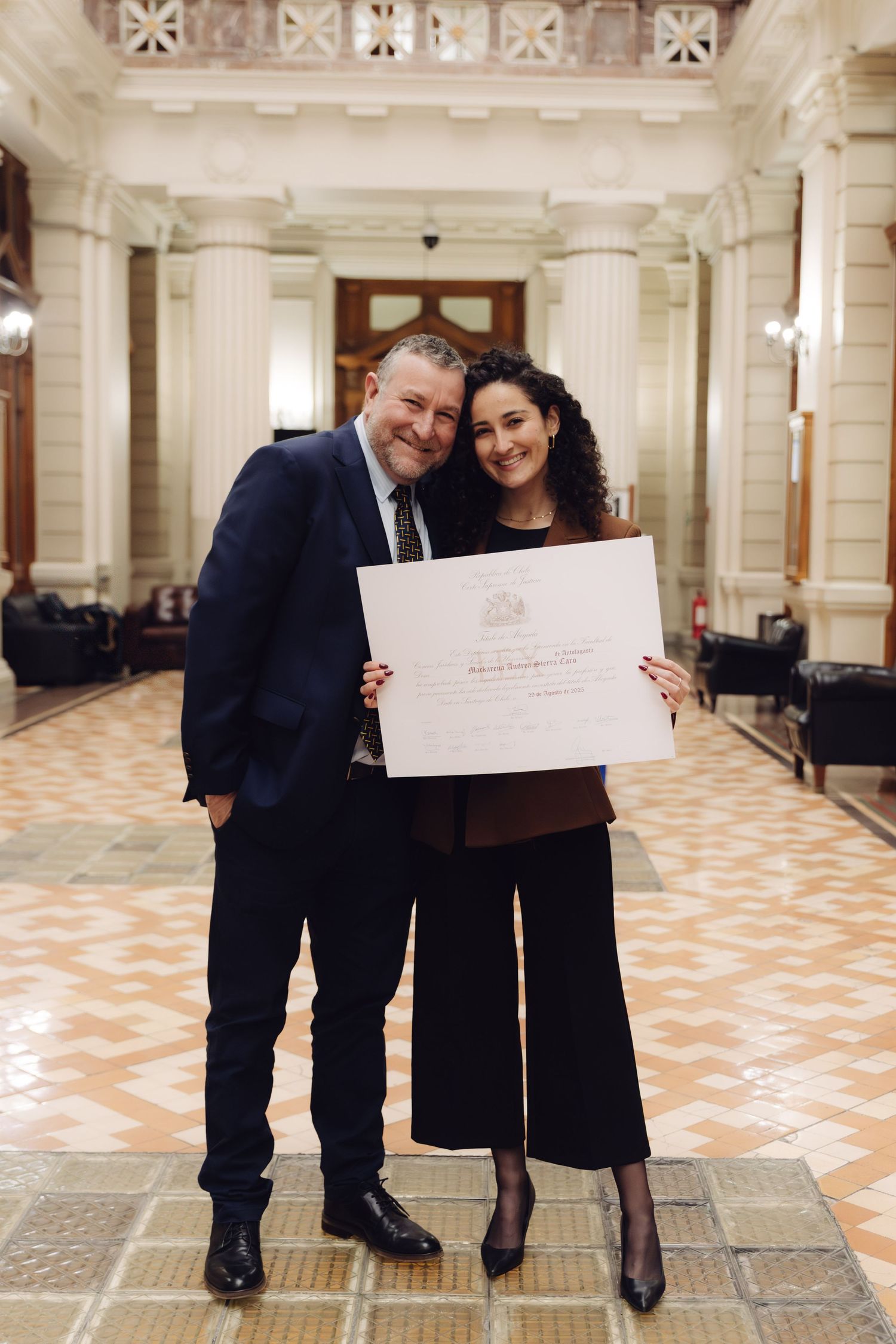 Man and woman proudly hold a framed certificate in a grand hall.