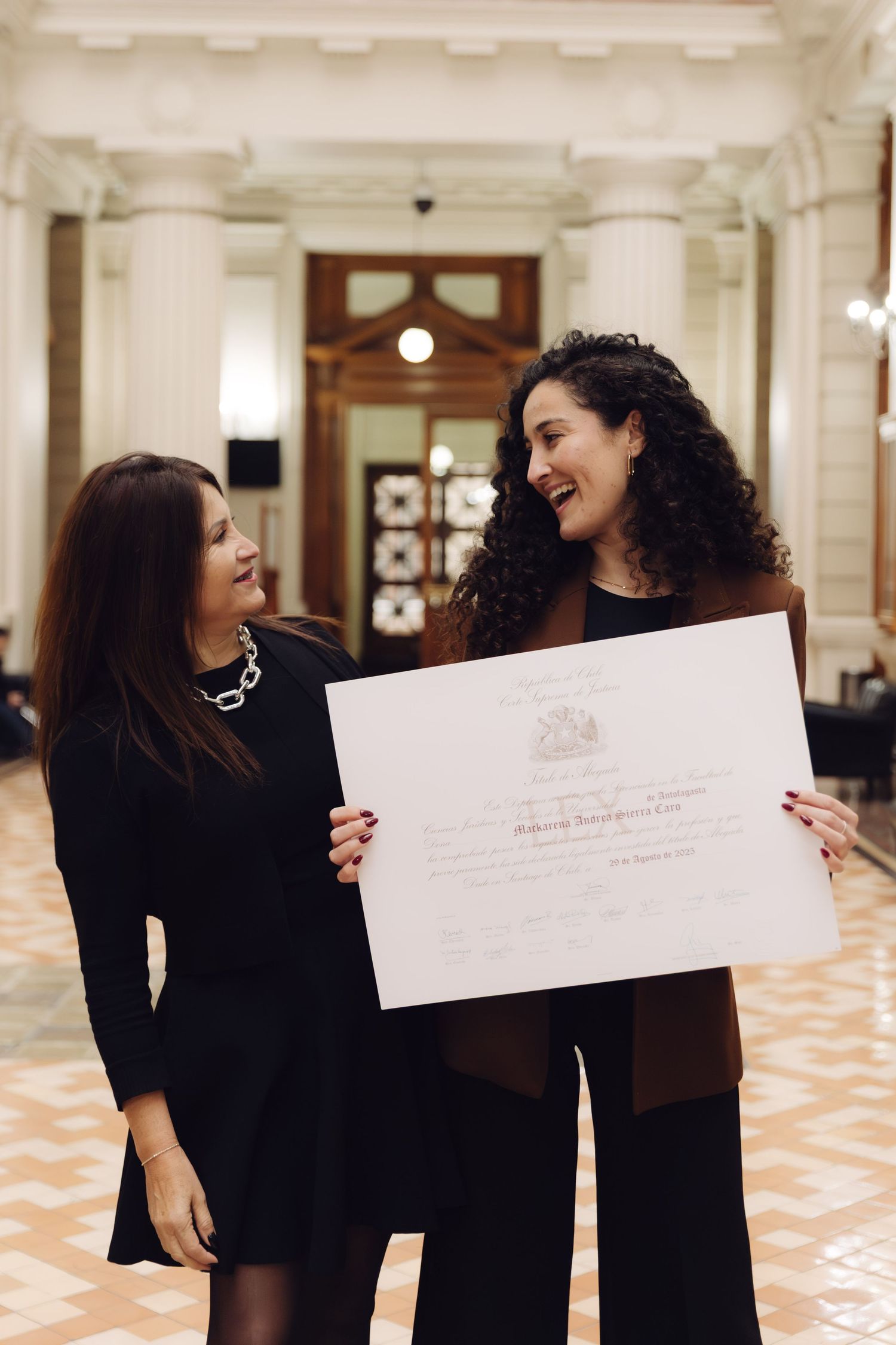 Two women laugh while holding an official certificate in a hall.