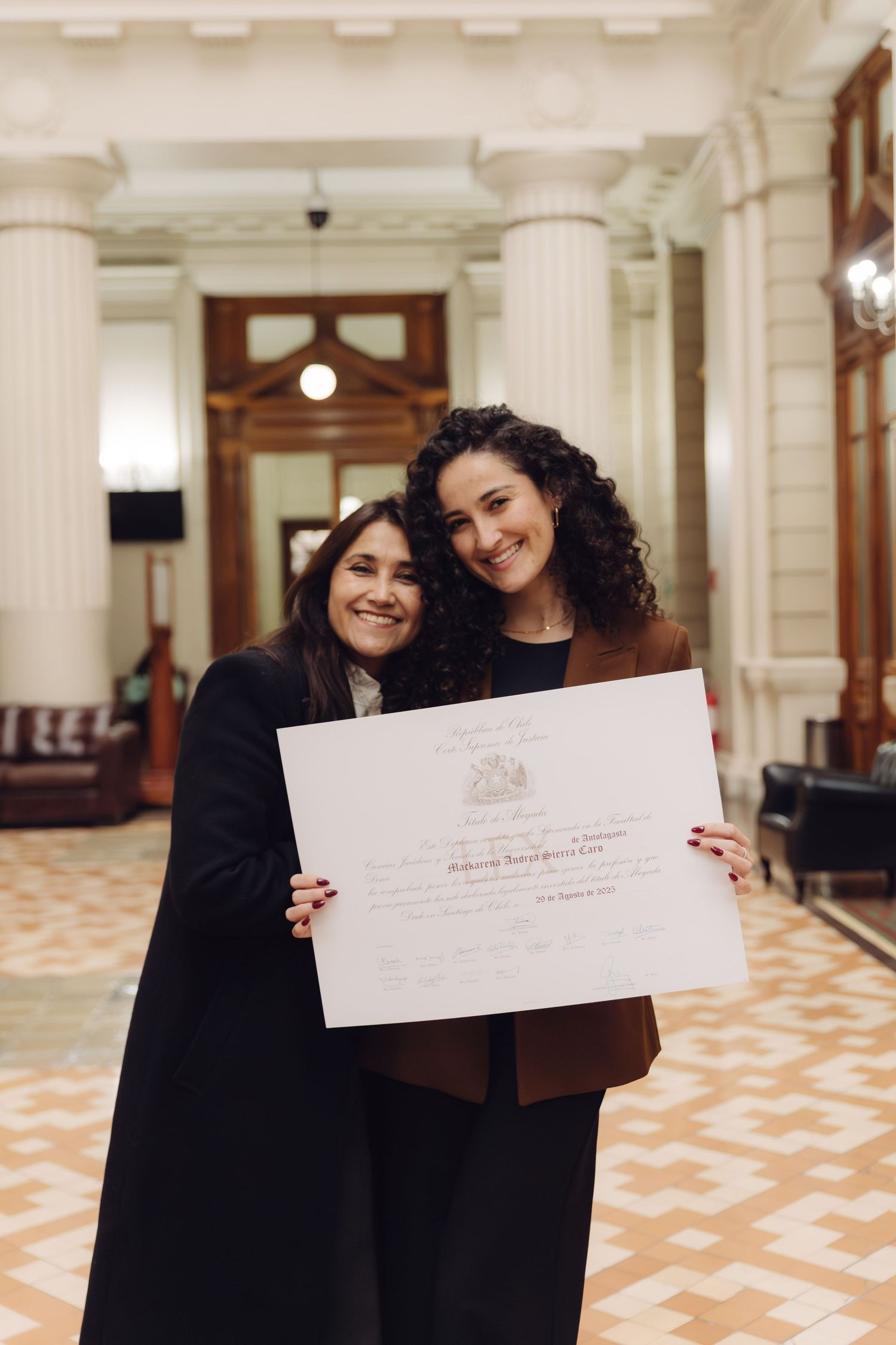 Two smiling women hold a large certificate in an elegant hall.