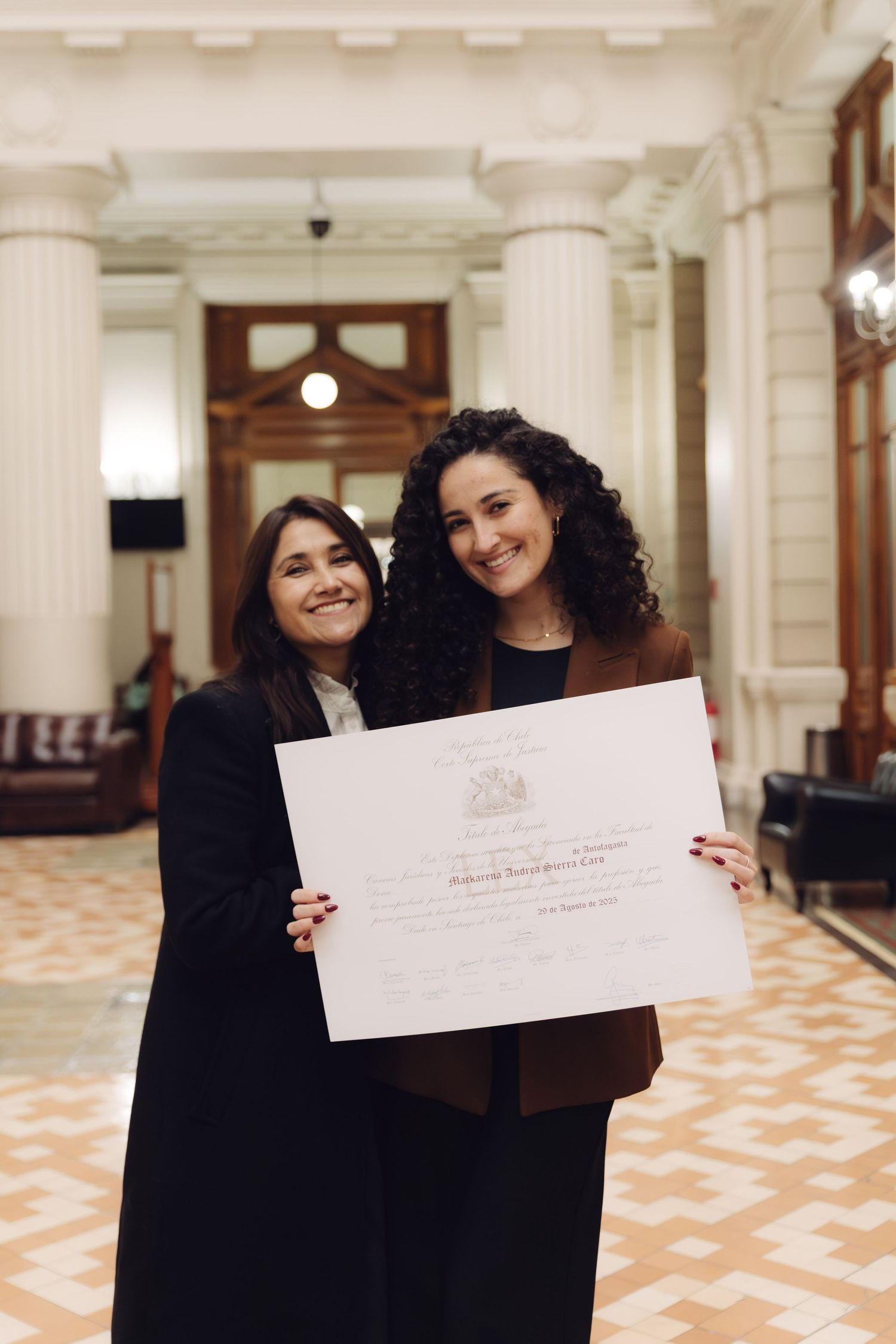 Two women proudly display an official certificate in a hallway.