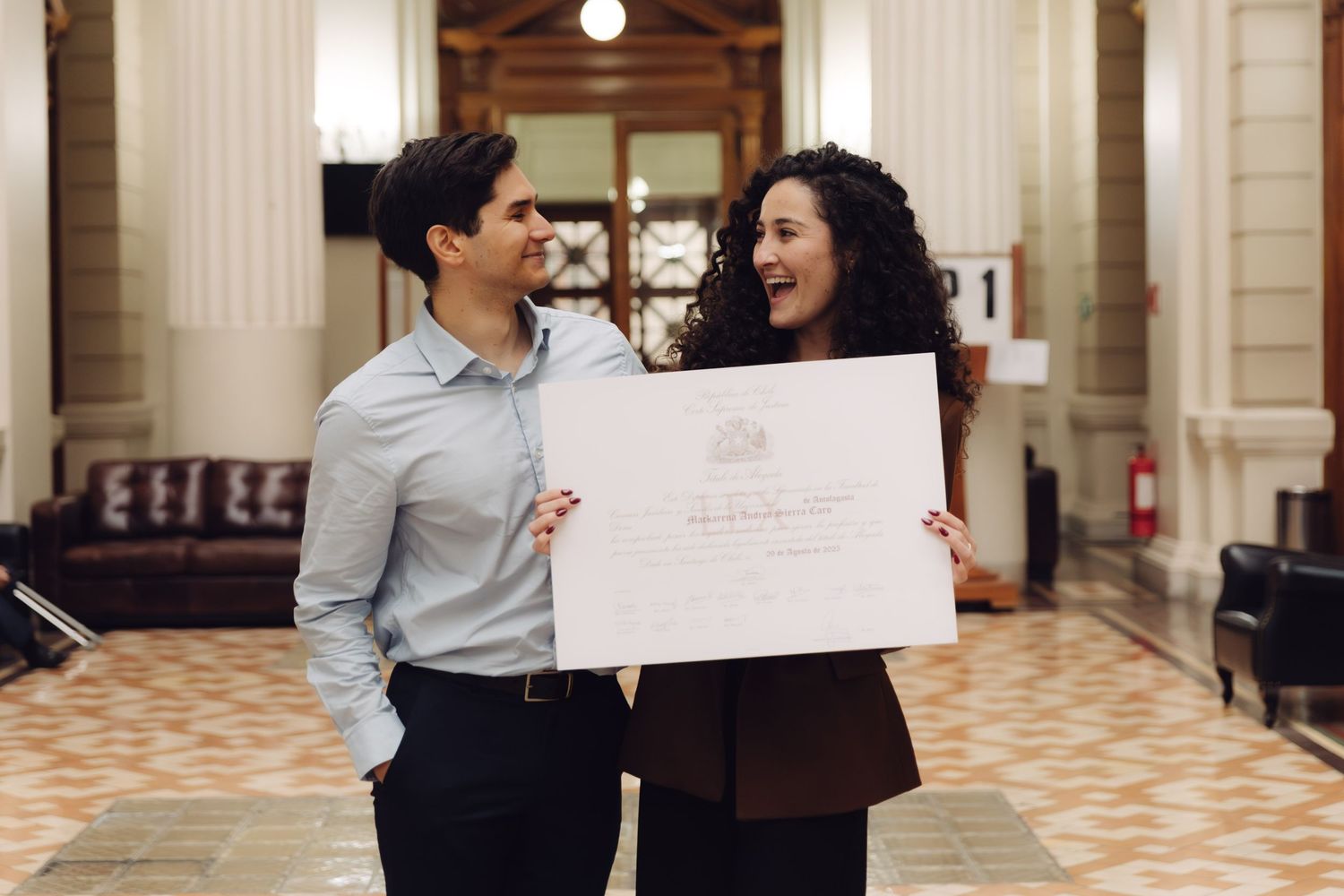 Couple smiling at each other while holding a graduation diploma in a grand hall.