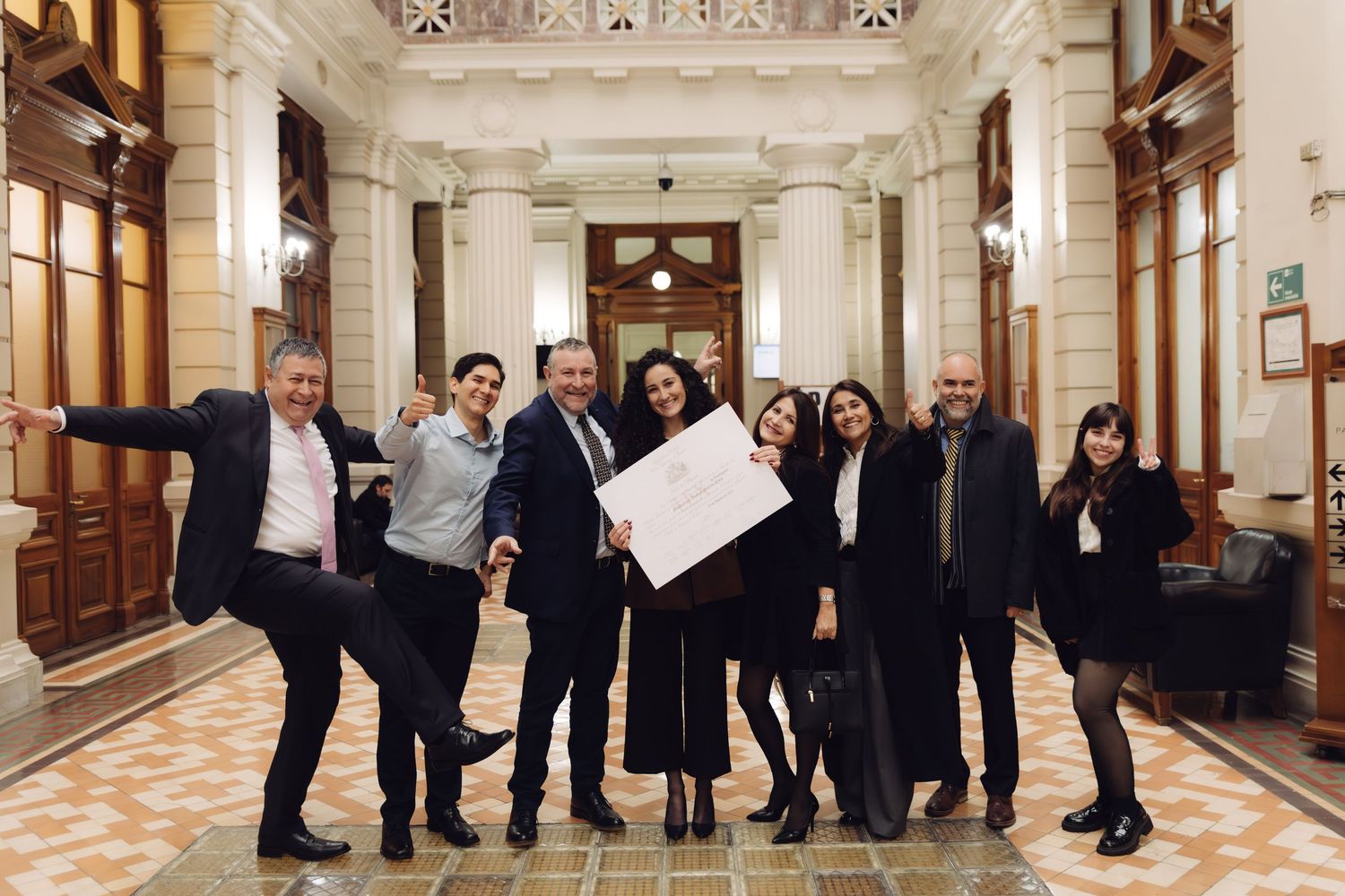 Joyful group of eight celebrating with a diploma in a classic building hallway.