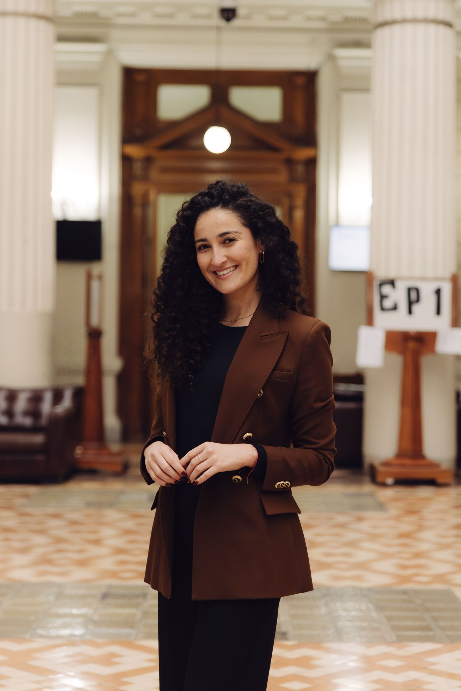 Smiling woman in brown blazer posing in an elegant hall with columns.