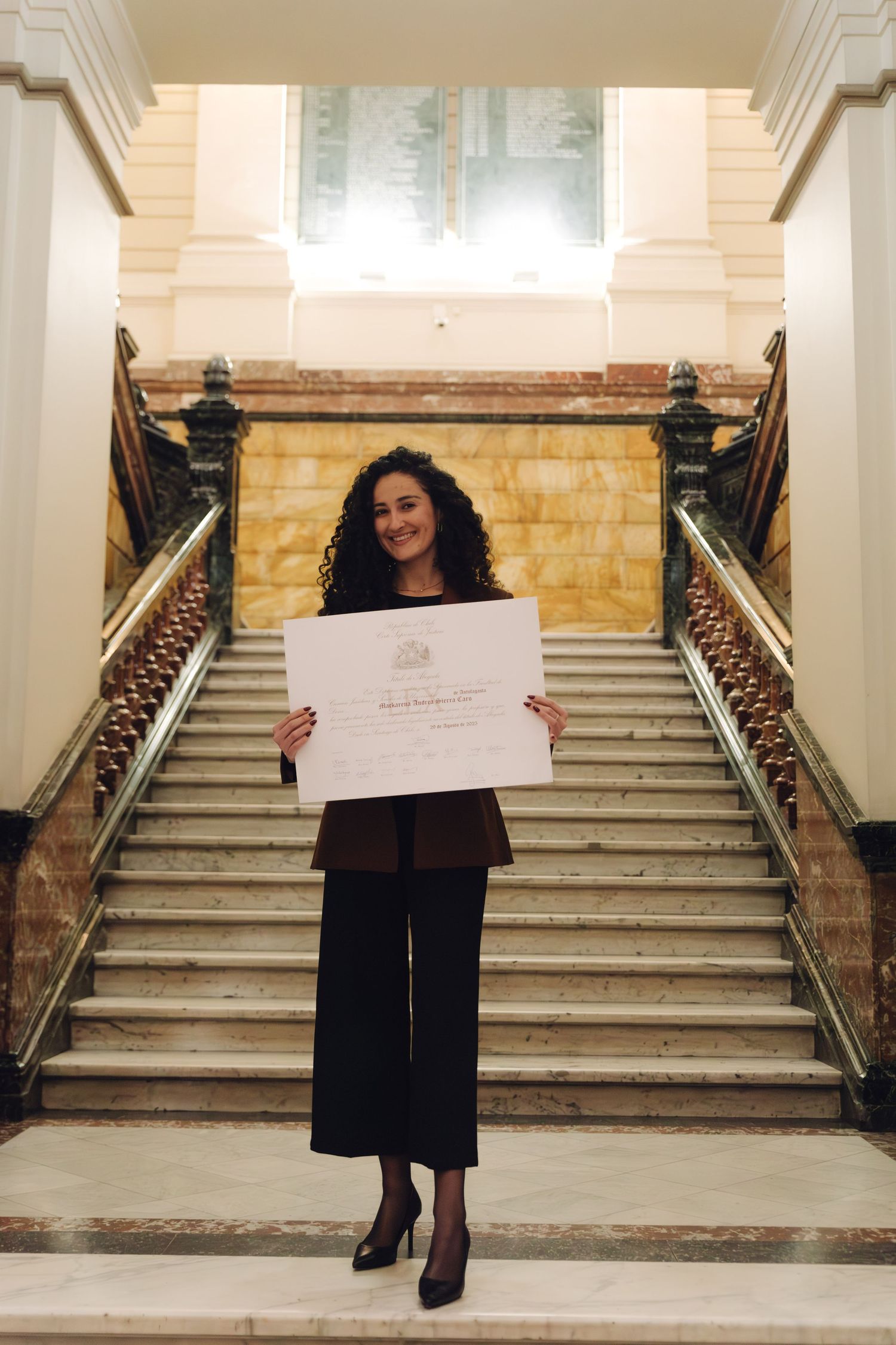 Woman in brown blazer holding a diploma at the base of marble stairs.