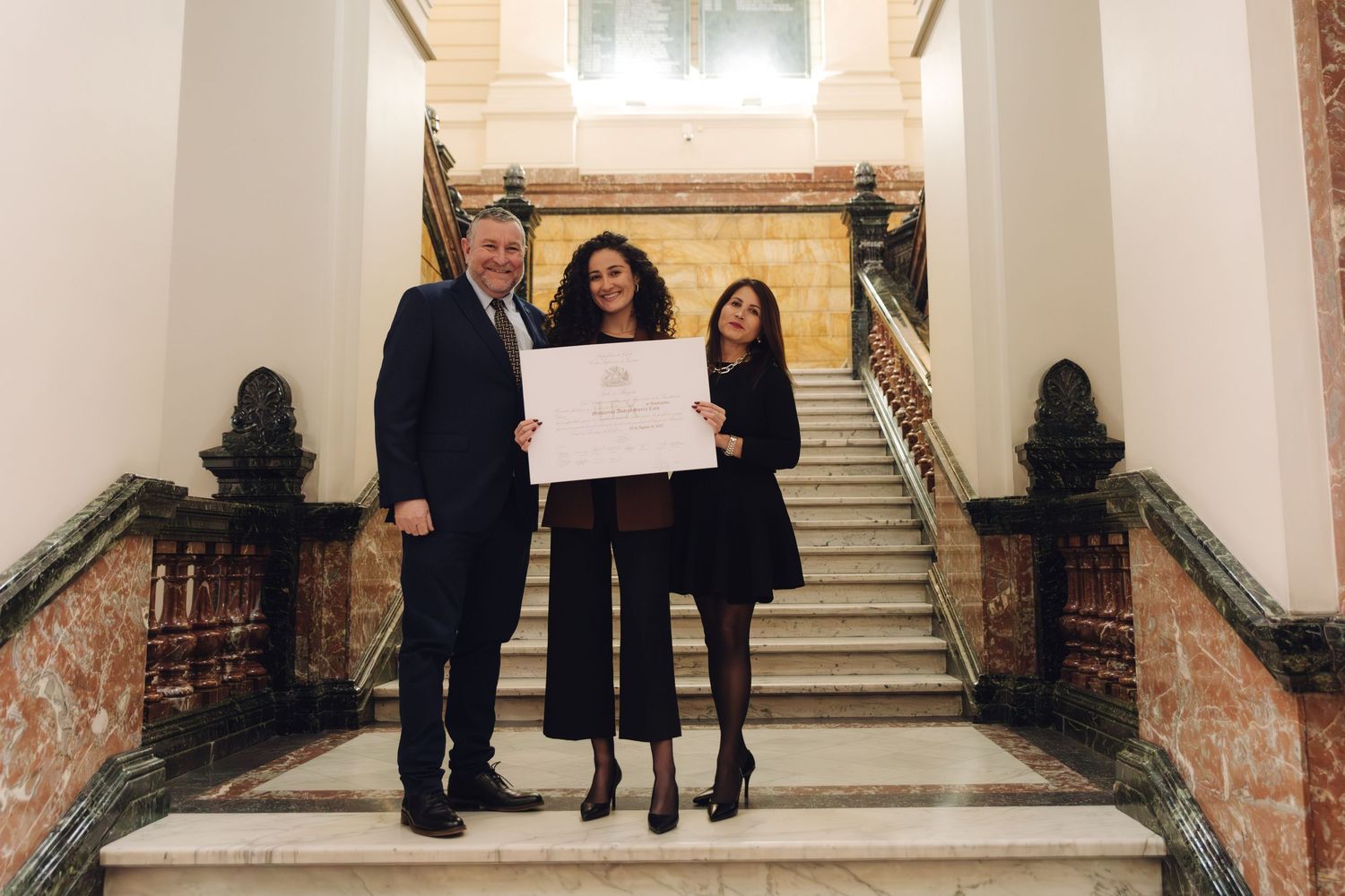 Three people posing together holding a diploma on an elegant staircase.