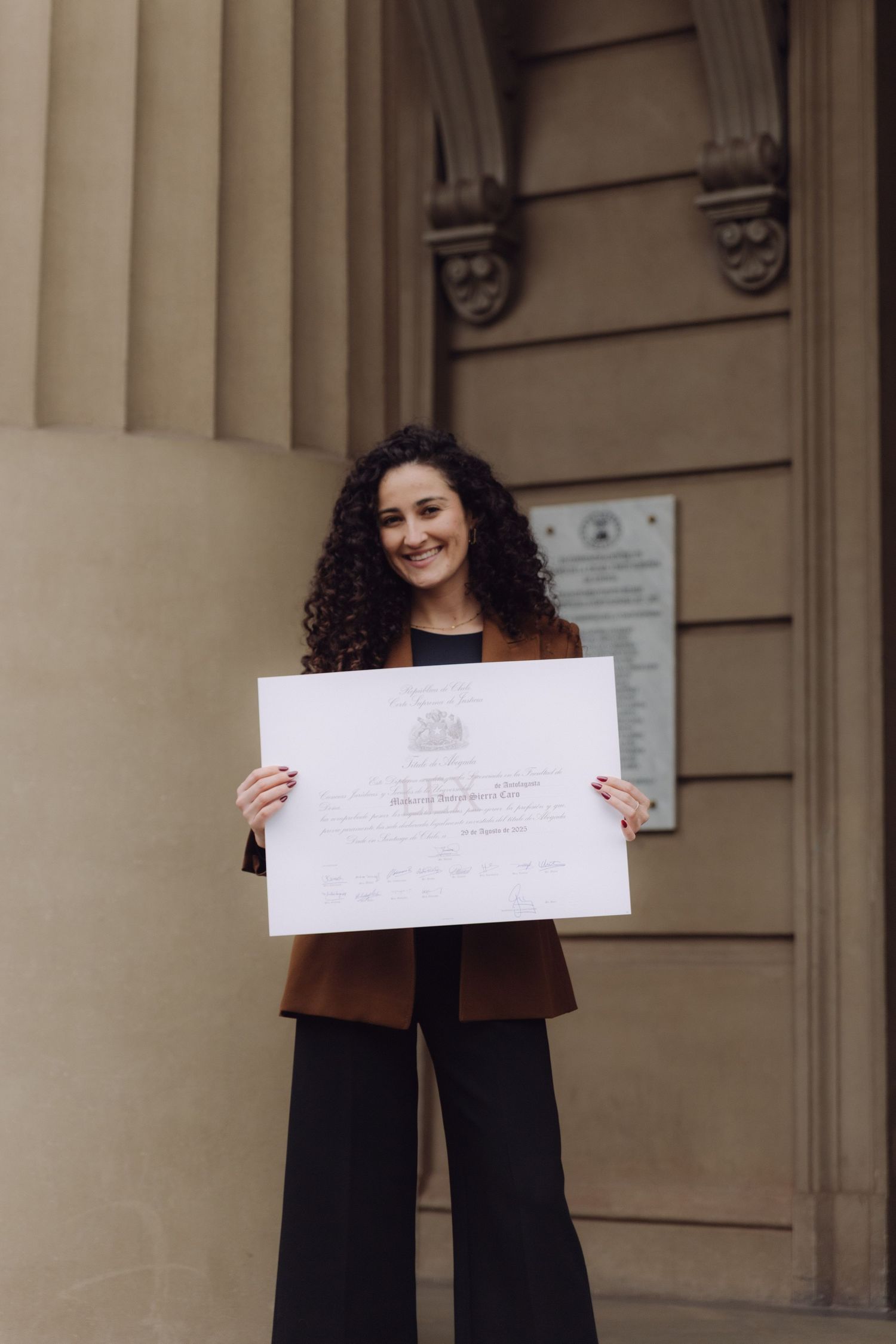 Young woman with curly hair proudly holding diploma certificate outside historic building.