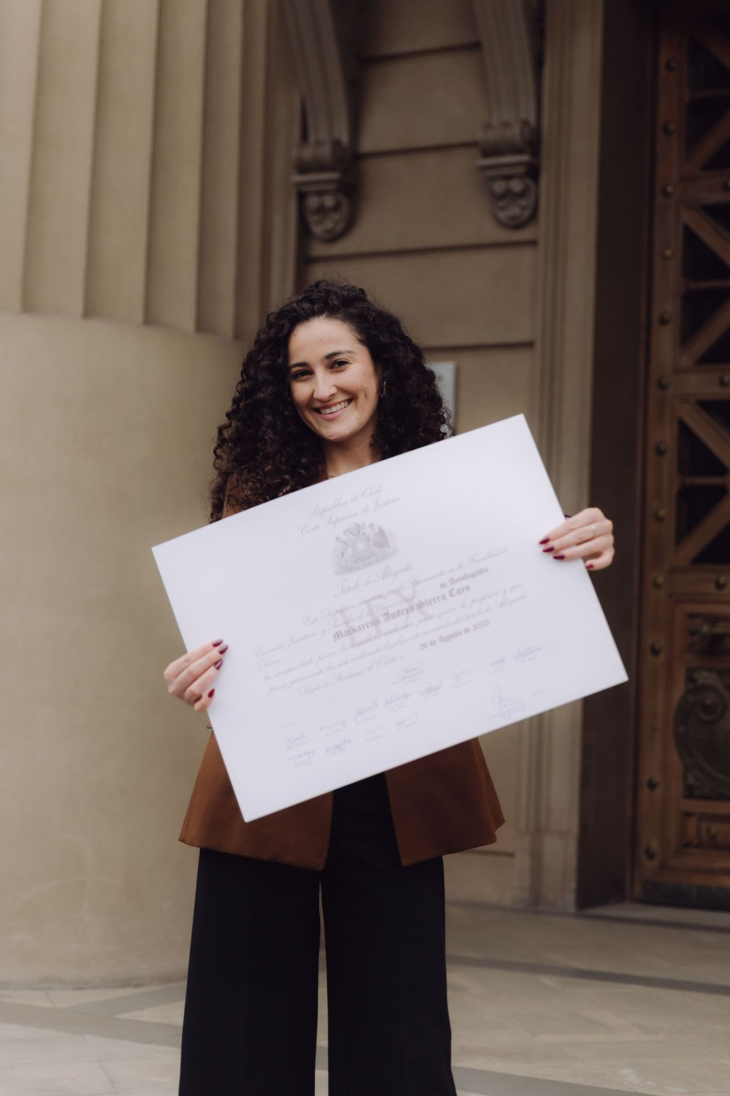 Smiling woman with curly hair displaying official diploma certificate outside classical building.