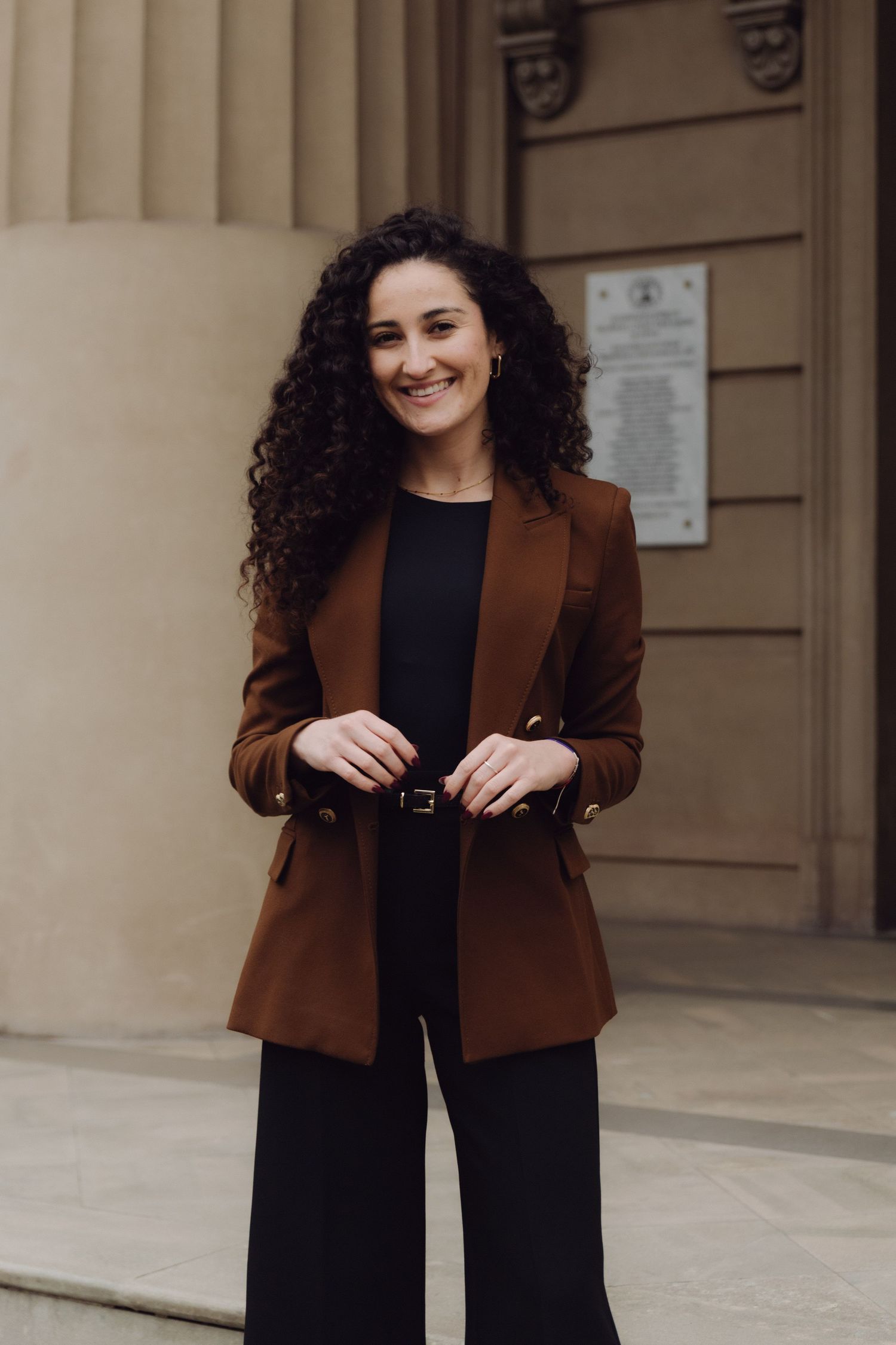 Professional woman in brown blazer and black outfit smiling outside historic stone building.