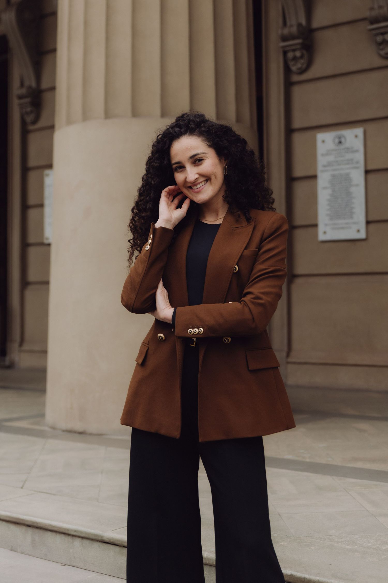 Elegant woman in brown blazer with curly hair posing outside classical architecture building.