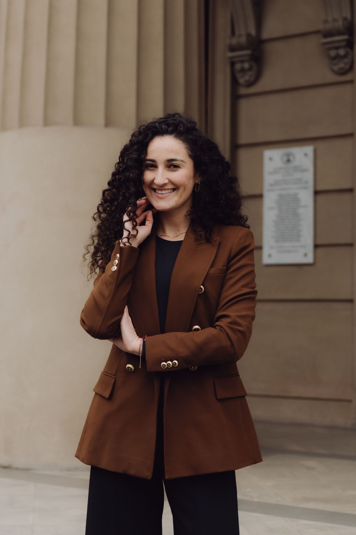 Cheerful woman in brown blazer with curly hair smiling outside historic building with plaque.