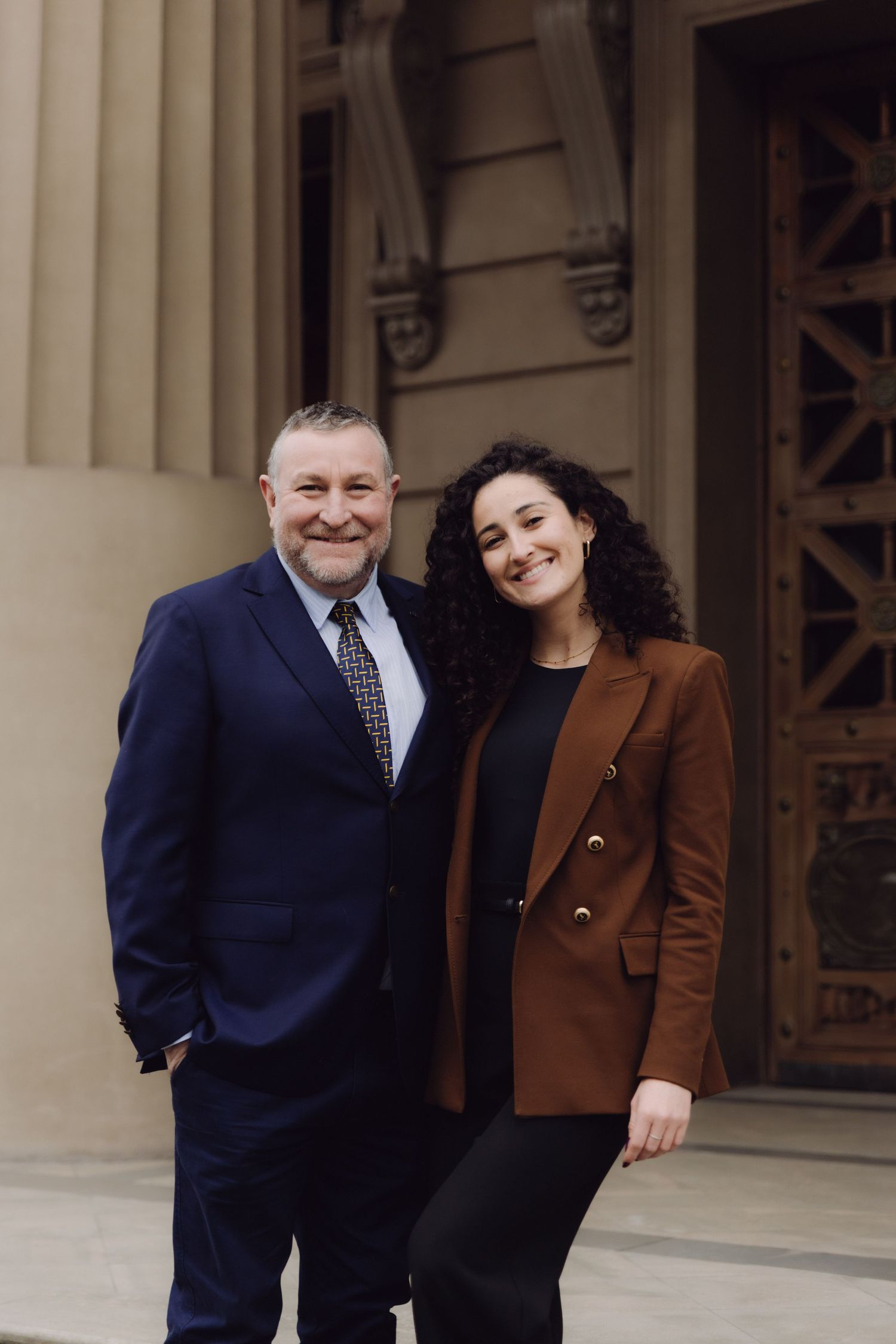 Man in navy suit and woman in brown blazer smiling together outside classical building.