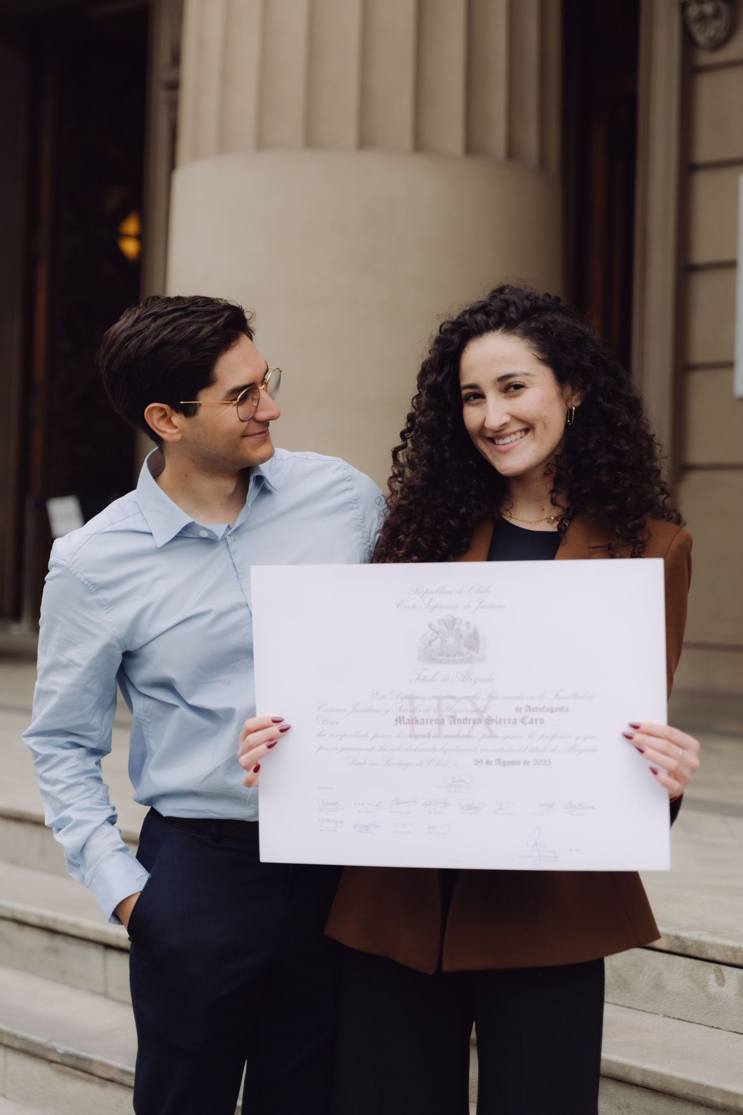 Young couple smiling together holding official diploma certificate on steps of historic building.