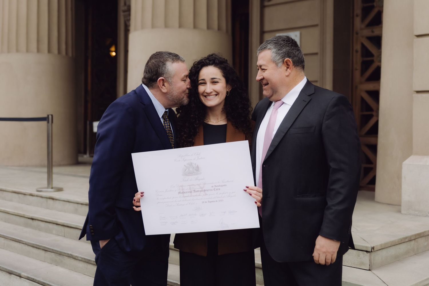 Woman proudly holds diploma flanked by two men in suits on building steps.