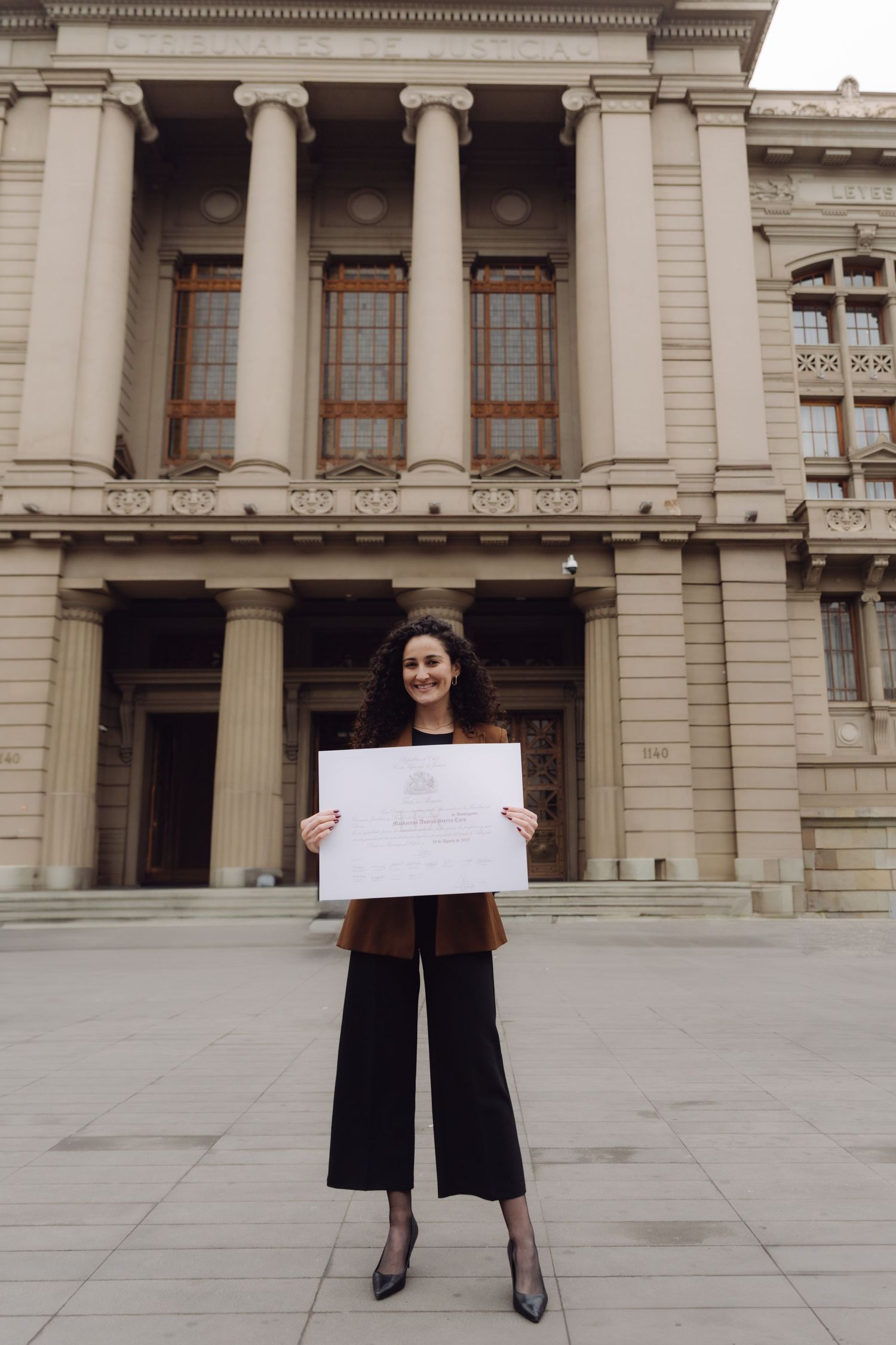 Graduate holds large diploma certificate in front of neoclassical building columns.