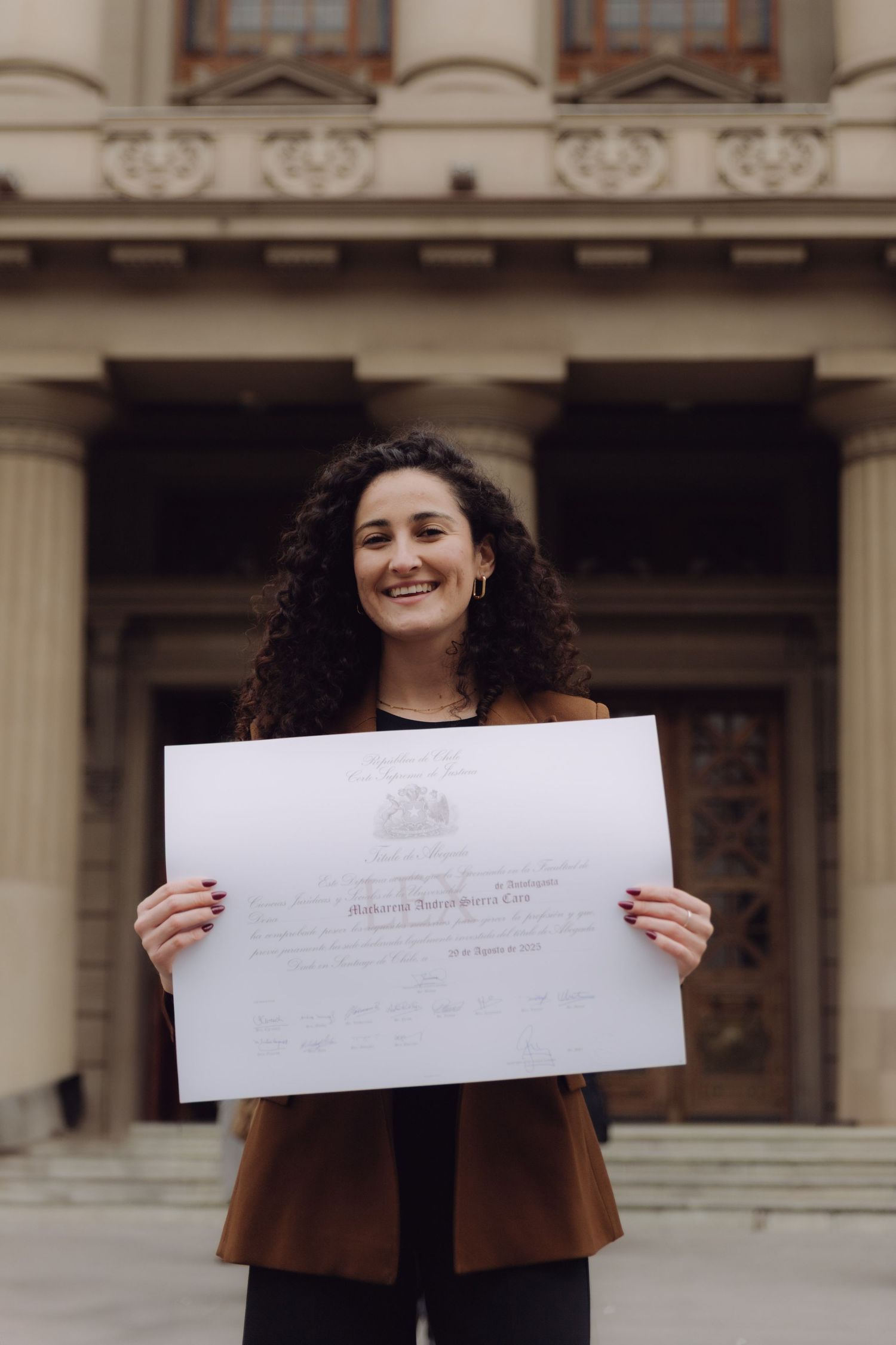 Smiling curly-haired woman holds official certificate outside historic building.