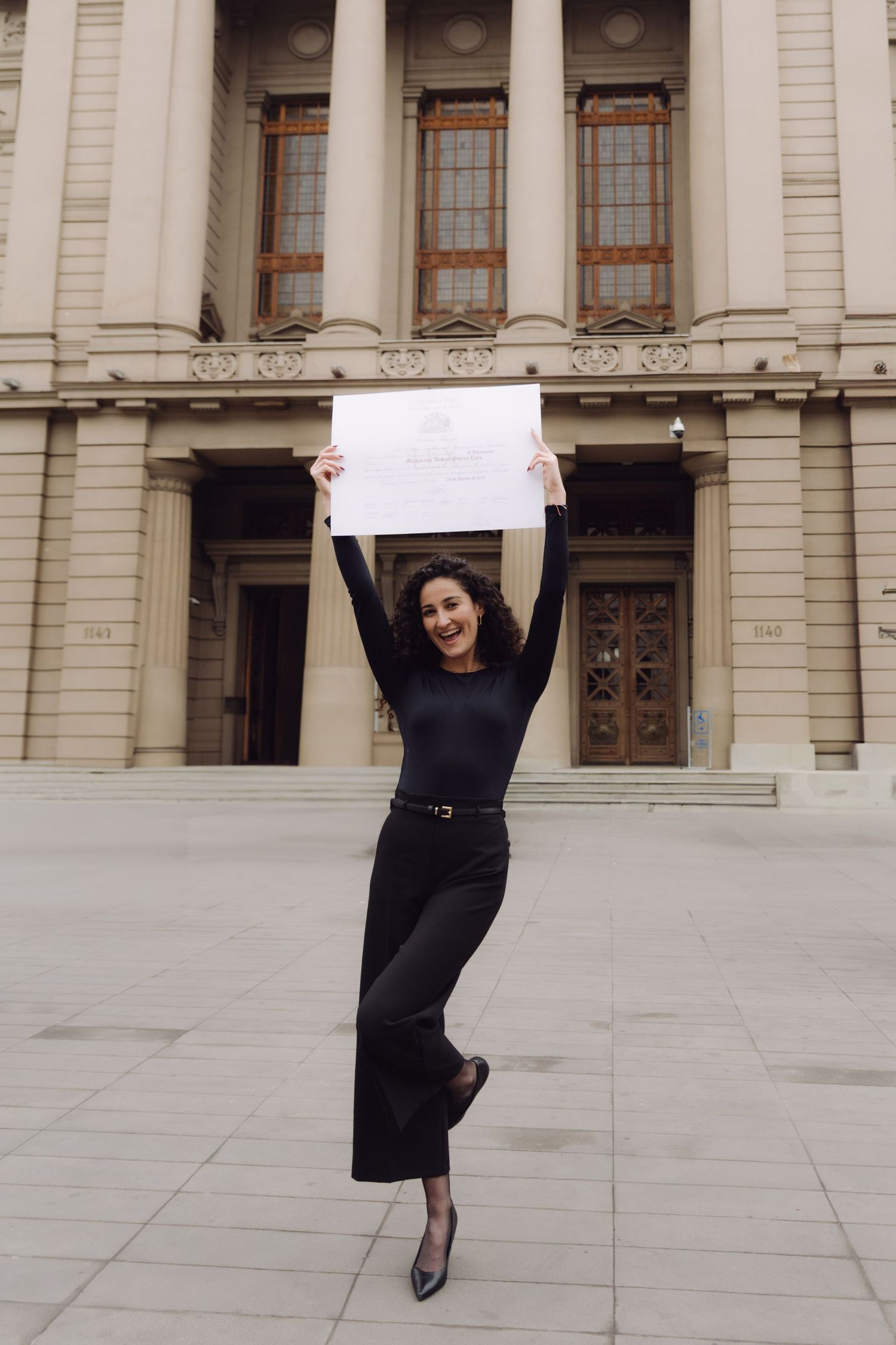 Joyful woman raises diploma overhead celebrating graduation outside grand building.
