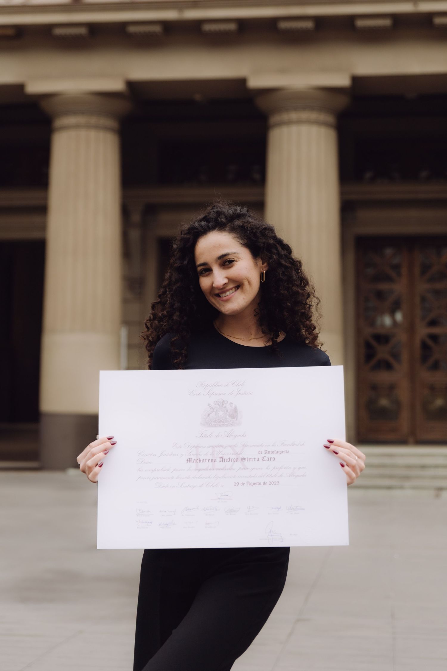 Proud graduate with curly hair displays official certificate outside columned building.
