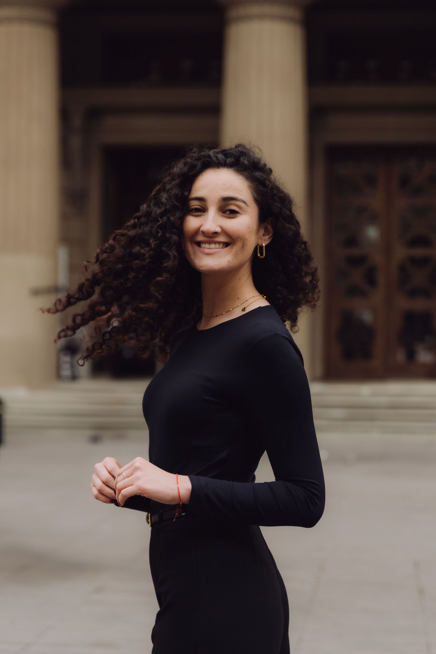 Smiling woman with curly hair in black dress poses outside historic building.