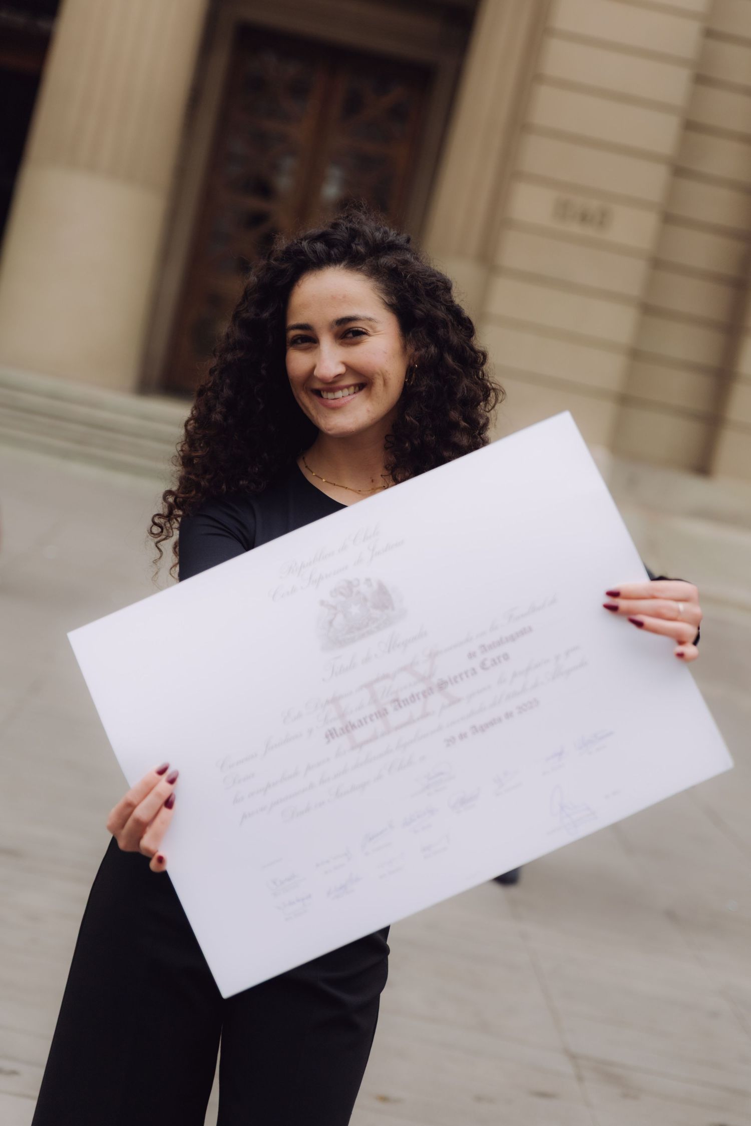 Happy curly-haired graduate proudly holds large official diploma certificate outdoors.