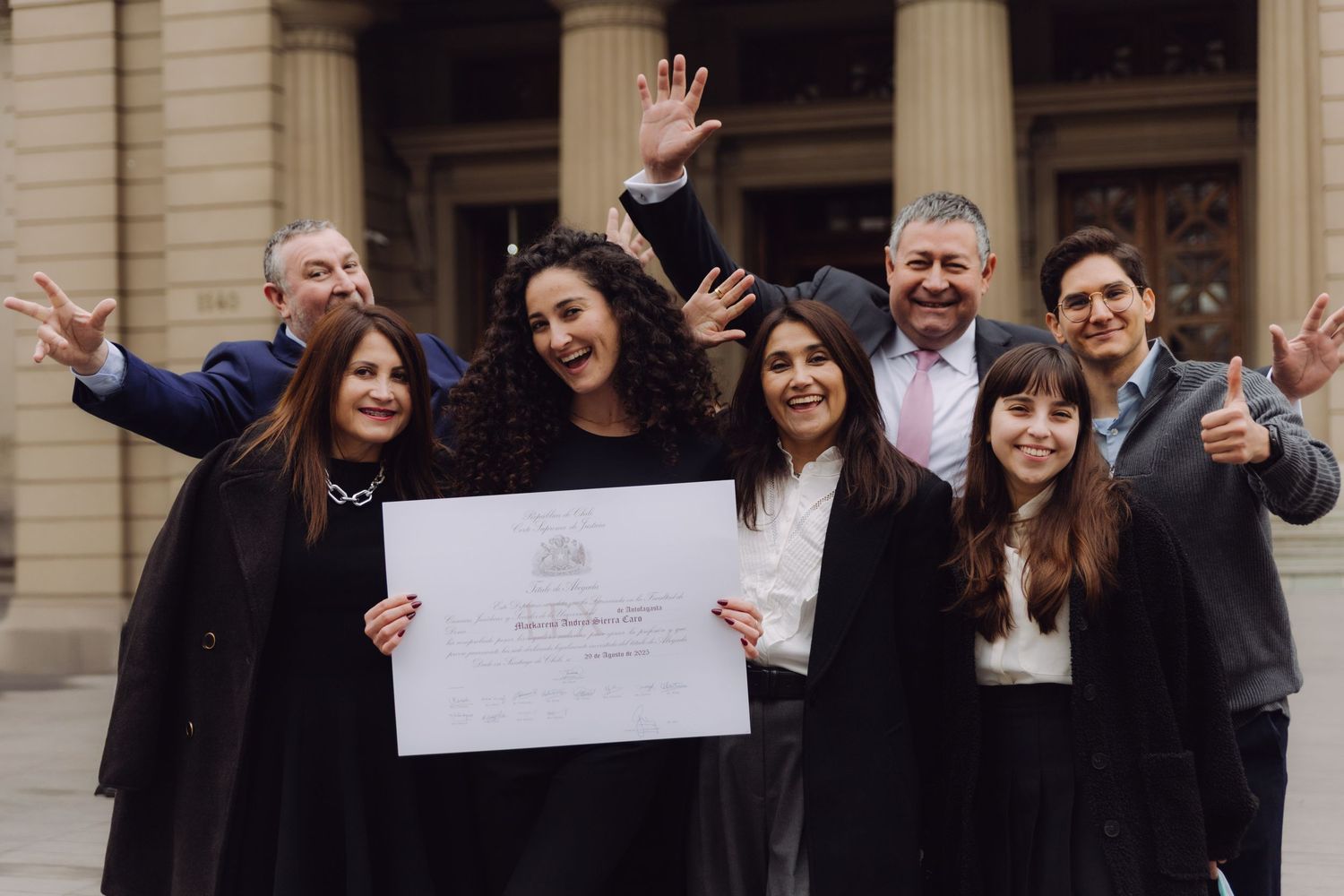 Excited group of seven people celebrate graduation holding diploma outside building.