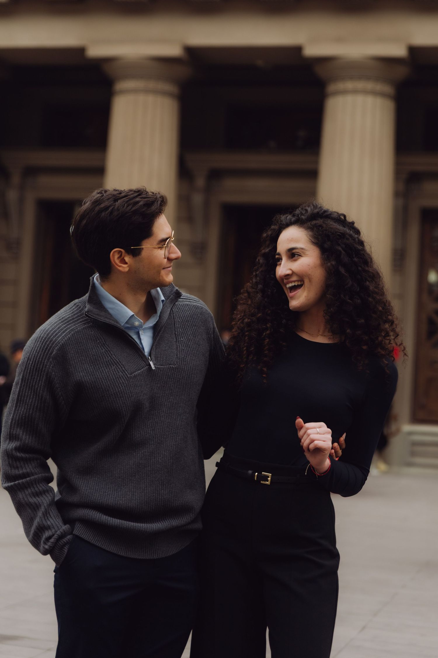 Young man and curly-haired woman laugh together outside neoclassical building.
