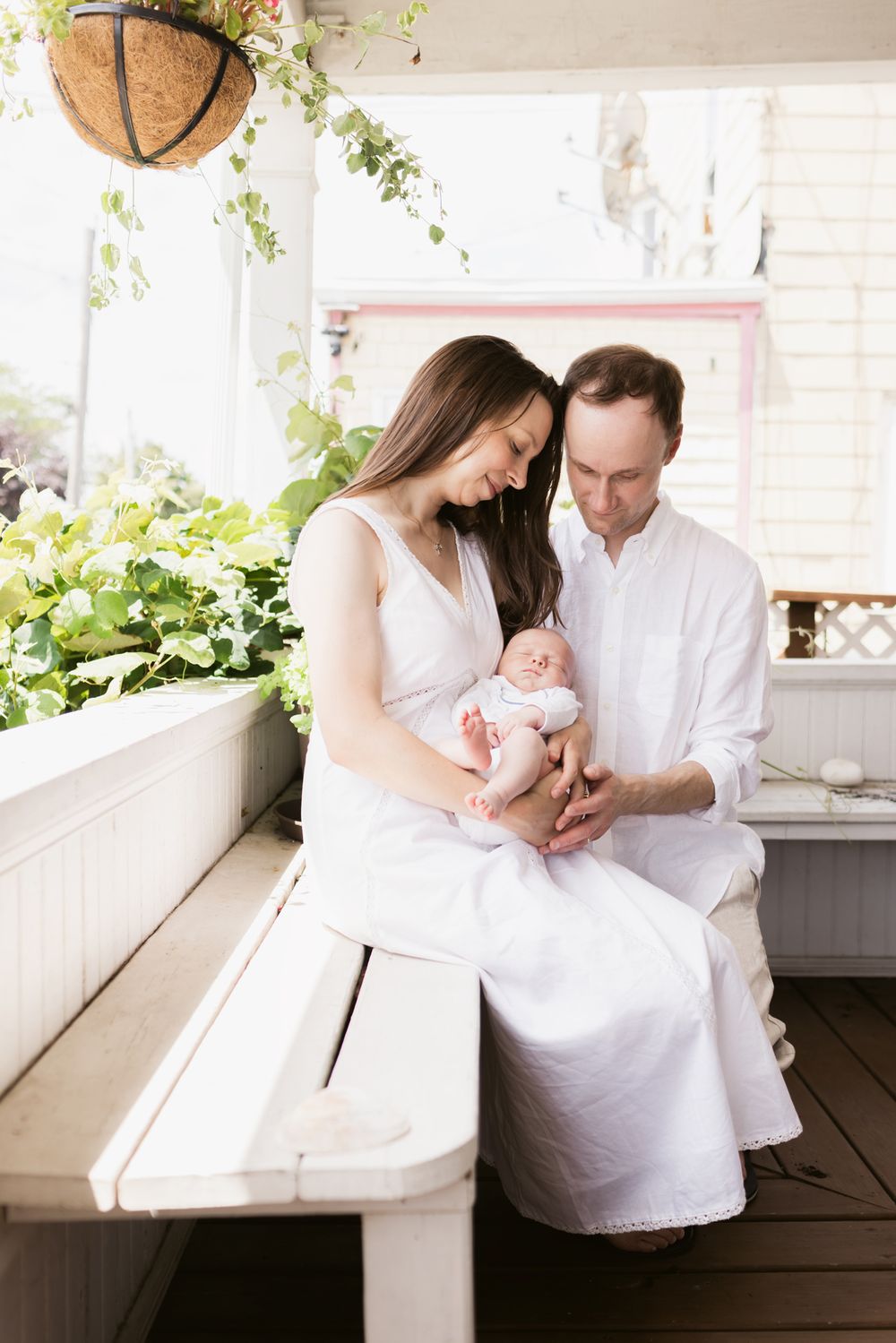 Mother and father wearing white sit on a white porch bench with plants around them holding a newborn baby in Winthrop, Massachusetts.