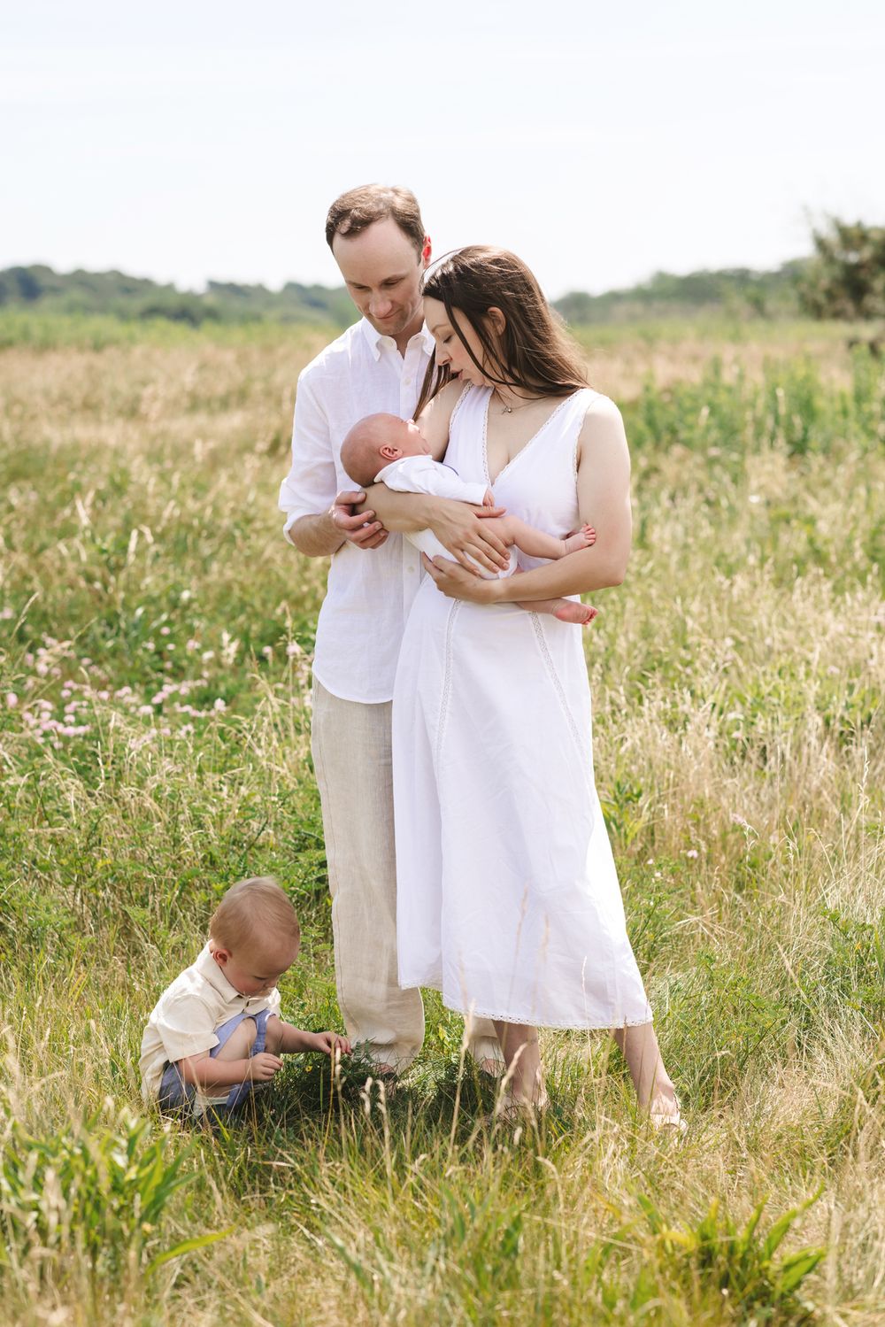 A family poses in a grassy field as toddler plays at parents' feet during a newborn family shoot on a partly sunny day.