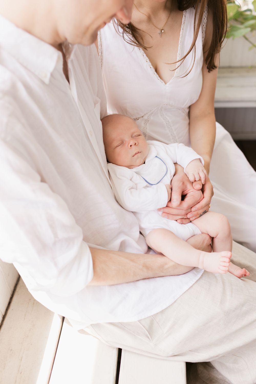 Couple wearing white snuggles baby boy on their lap while sitting on a bench on a porch for newborn photos. Manchester newborn photographer