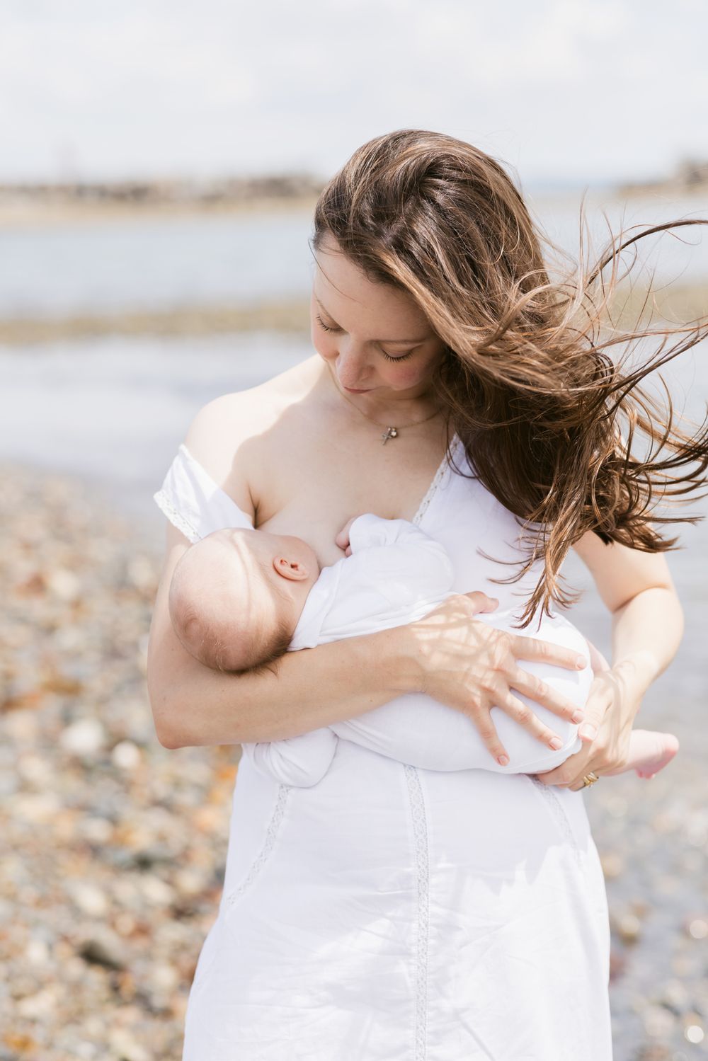 A mother wearing white dress with long dark hair blowing in the wind breastfeeds newborn while standing on rocky beach for newborn photos.