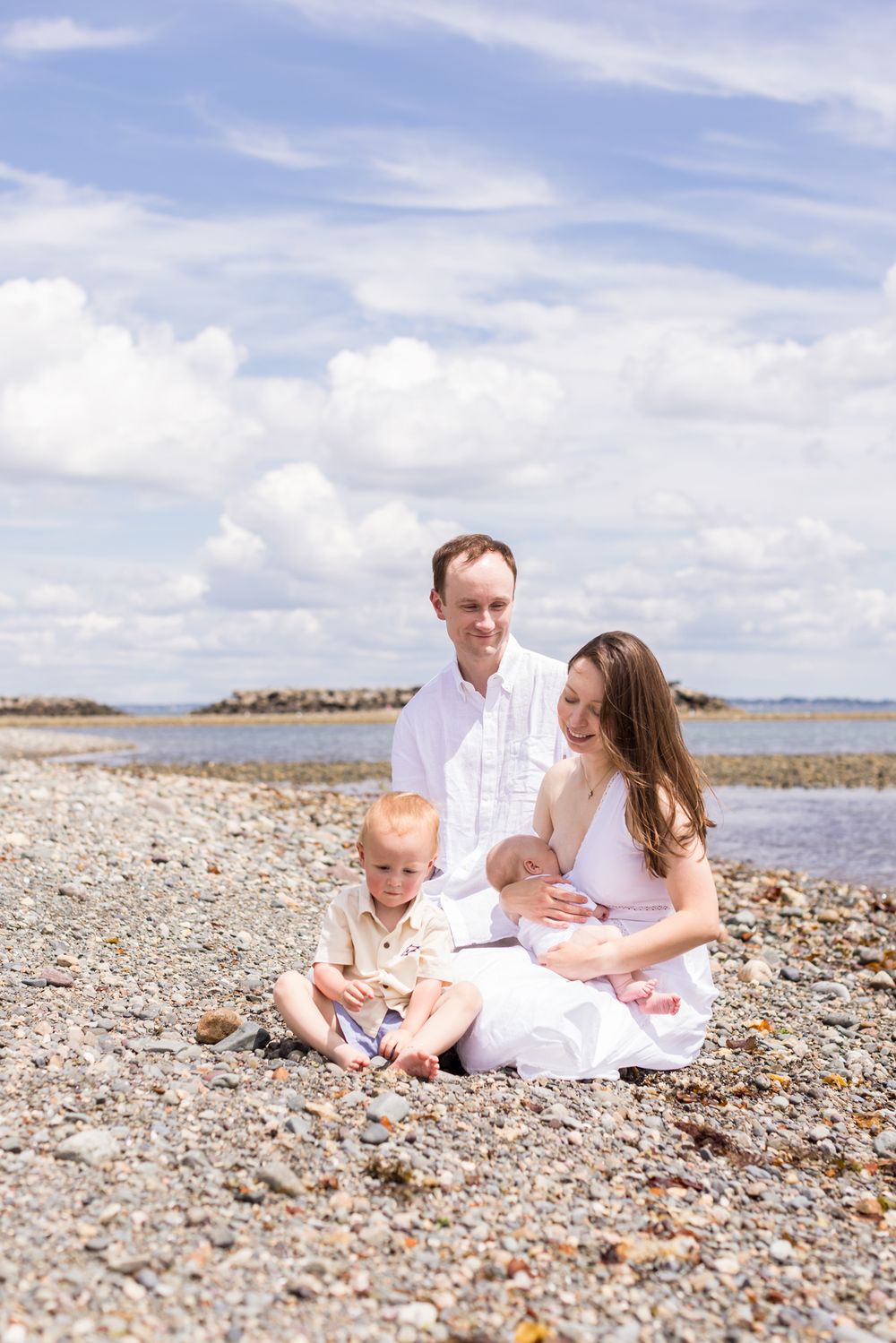 A smiling husband looks at wife smiling at toddler son while holding baby boy and sitting on a rocky beach under partly cloudy skies.
