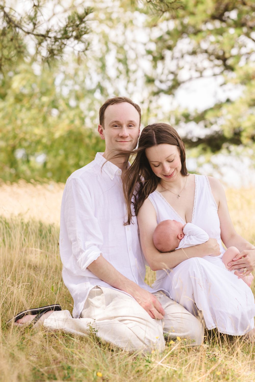 Mother looks down lovingly at newborn while siting next to husband in a grassy field for newborn photos. North Shore newborn photographer