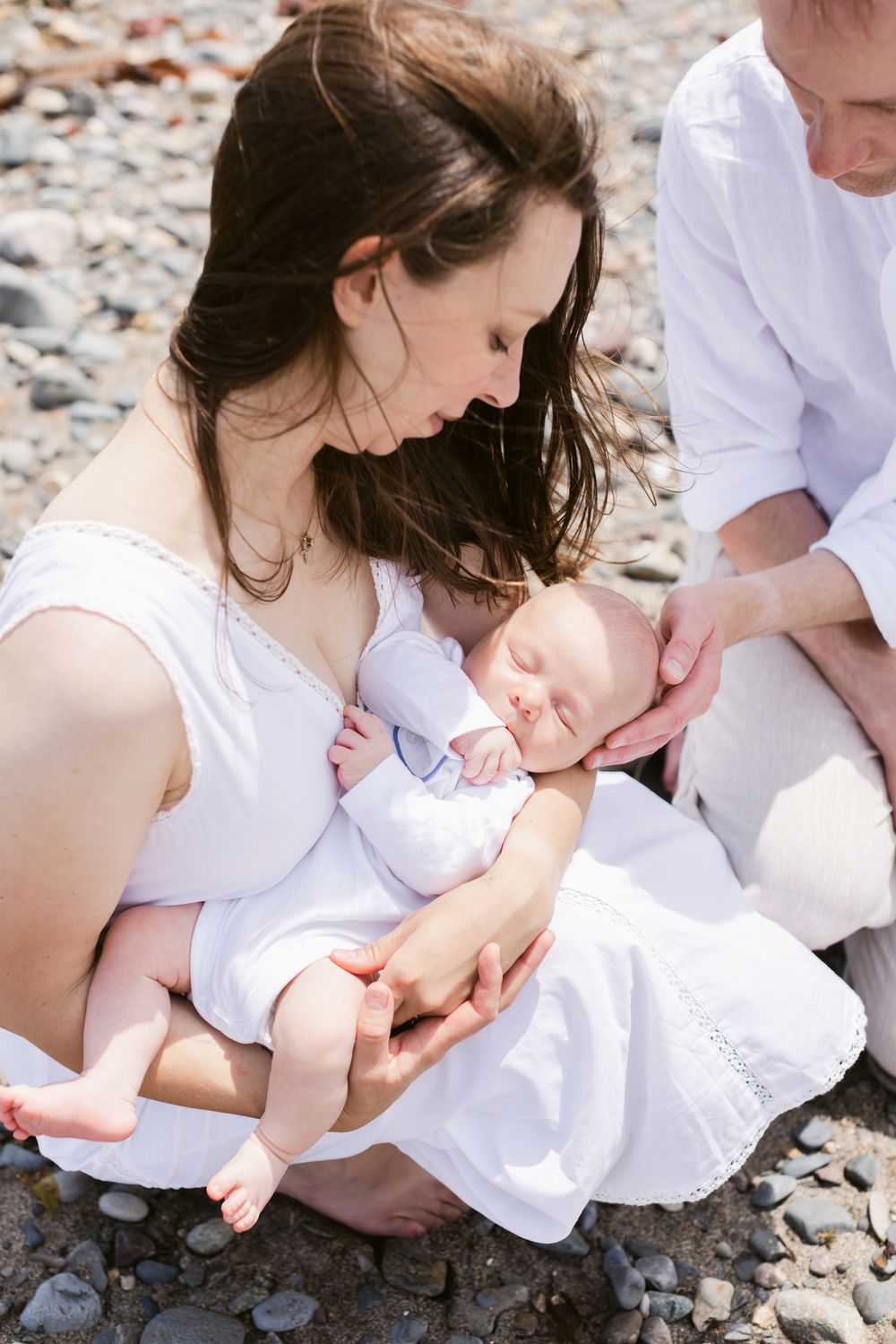 A mother and father wearing white kneel down holding a newborn son on a rocky beach for newborn photos. Southern Maine newborn photographer
