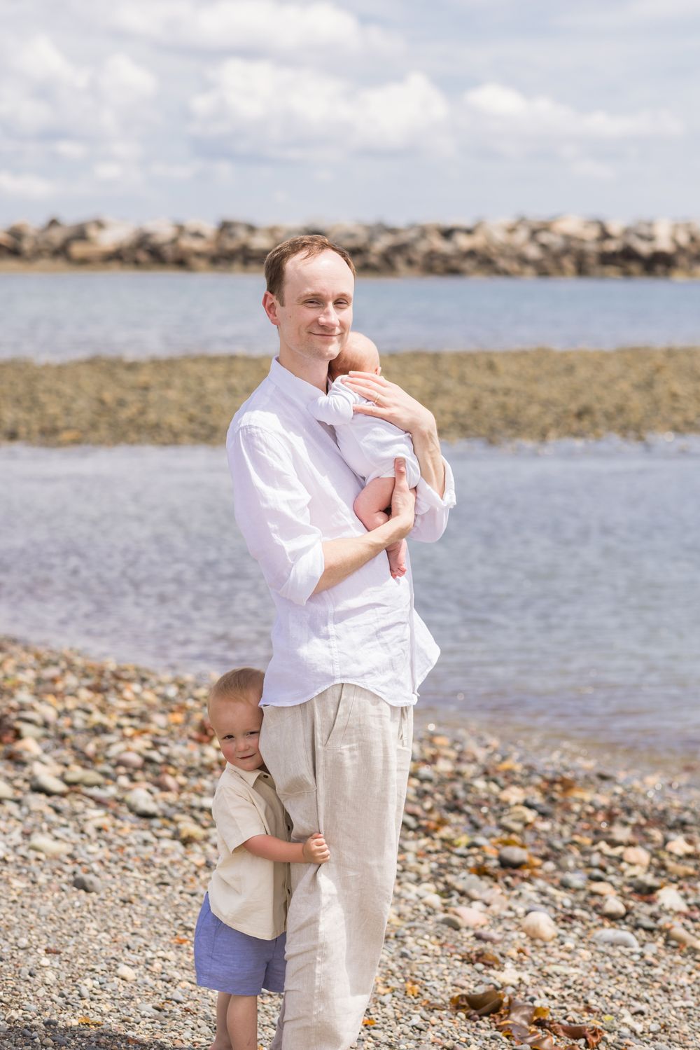 Father holds newborn son wearing white against his chest as toddler son holds his legs on a rocky beach on a party cloudy, sunny day.