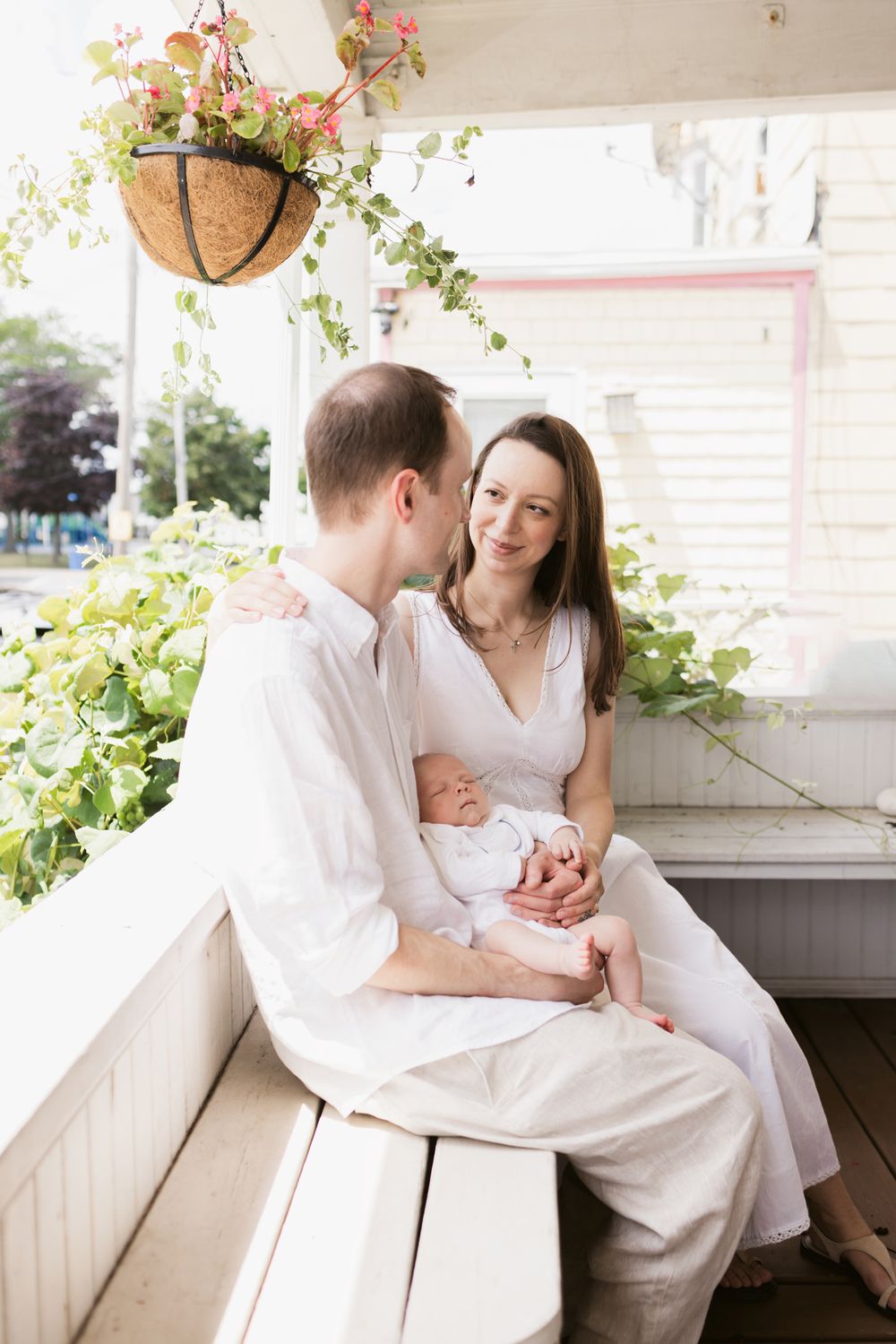 A couple wearing white gazes at each other while sitting on a white bench holding their newborn with plants around them in Winthrop, MA.