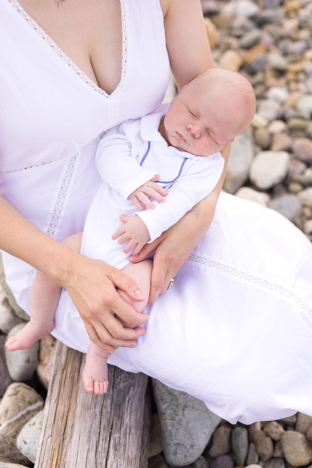 A mother wearing white dress embraces newborn wearing a white collared shirt while sitting on driftwood on coastal rocks for newborn photos