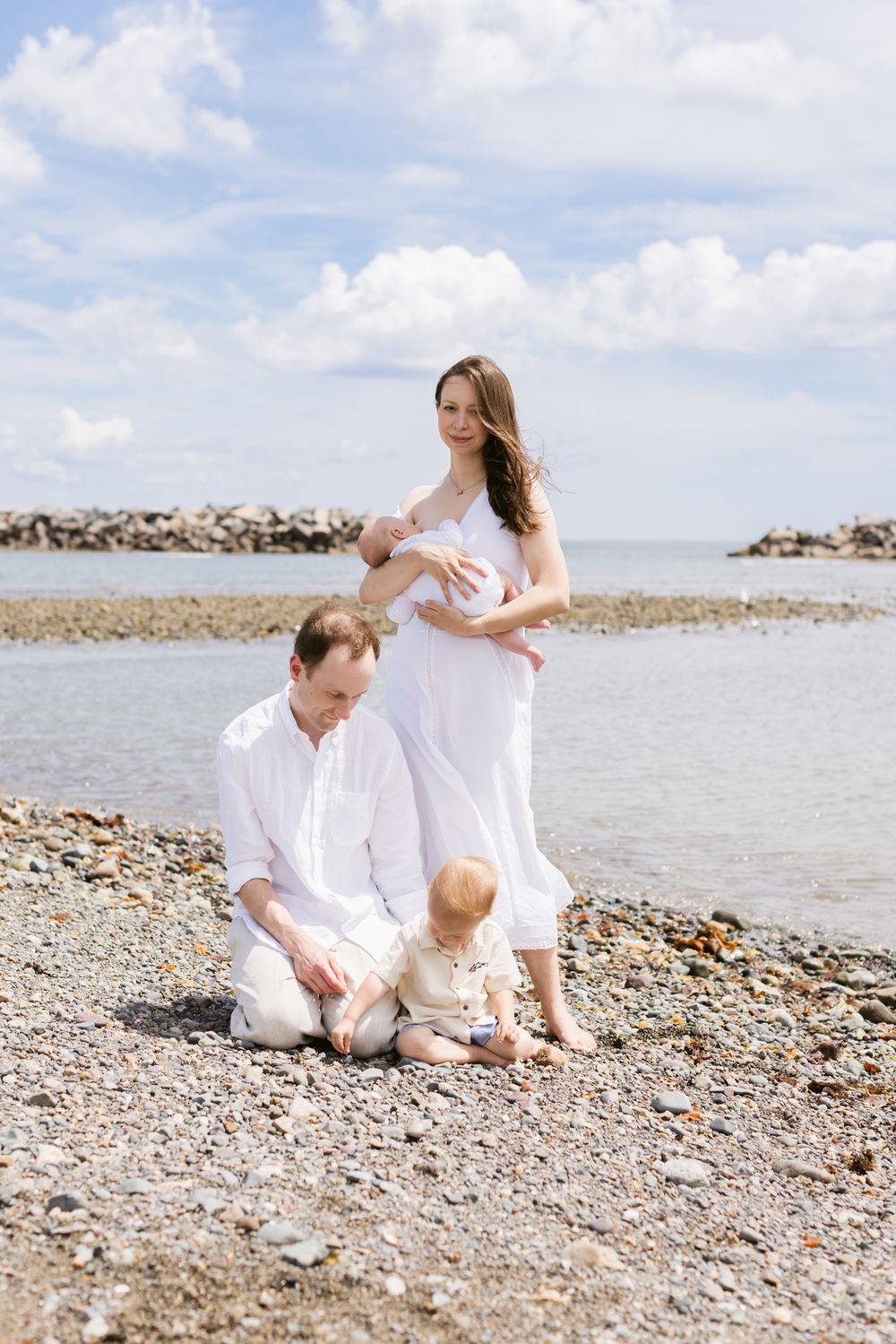 A mother wearing white dress holds newborn baby standing as husband and son play on rocky beach under partly cloudy skies for newborn photos