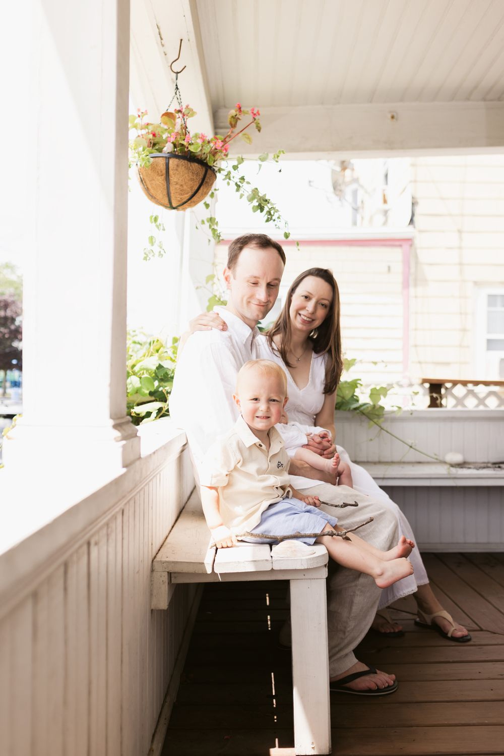 Happy family of four poses on a white porch bench, with hanging flower basket and green plants in background for family photos in Winthrop.