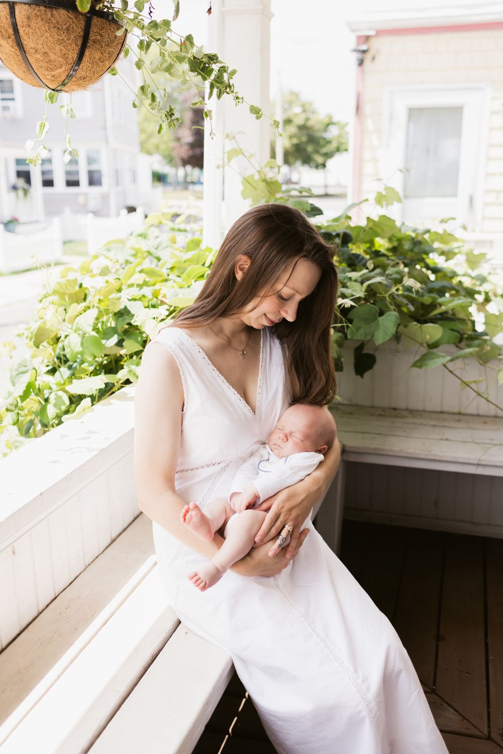 A young mother in white dress tenderly holds her newborn on a sunlit porch surrounded by green plants. Southern Maine newborn photographer.