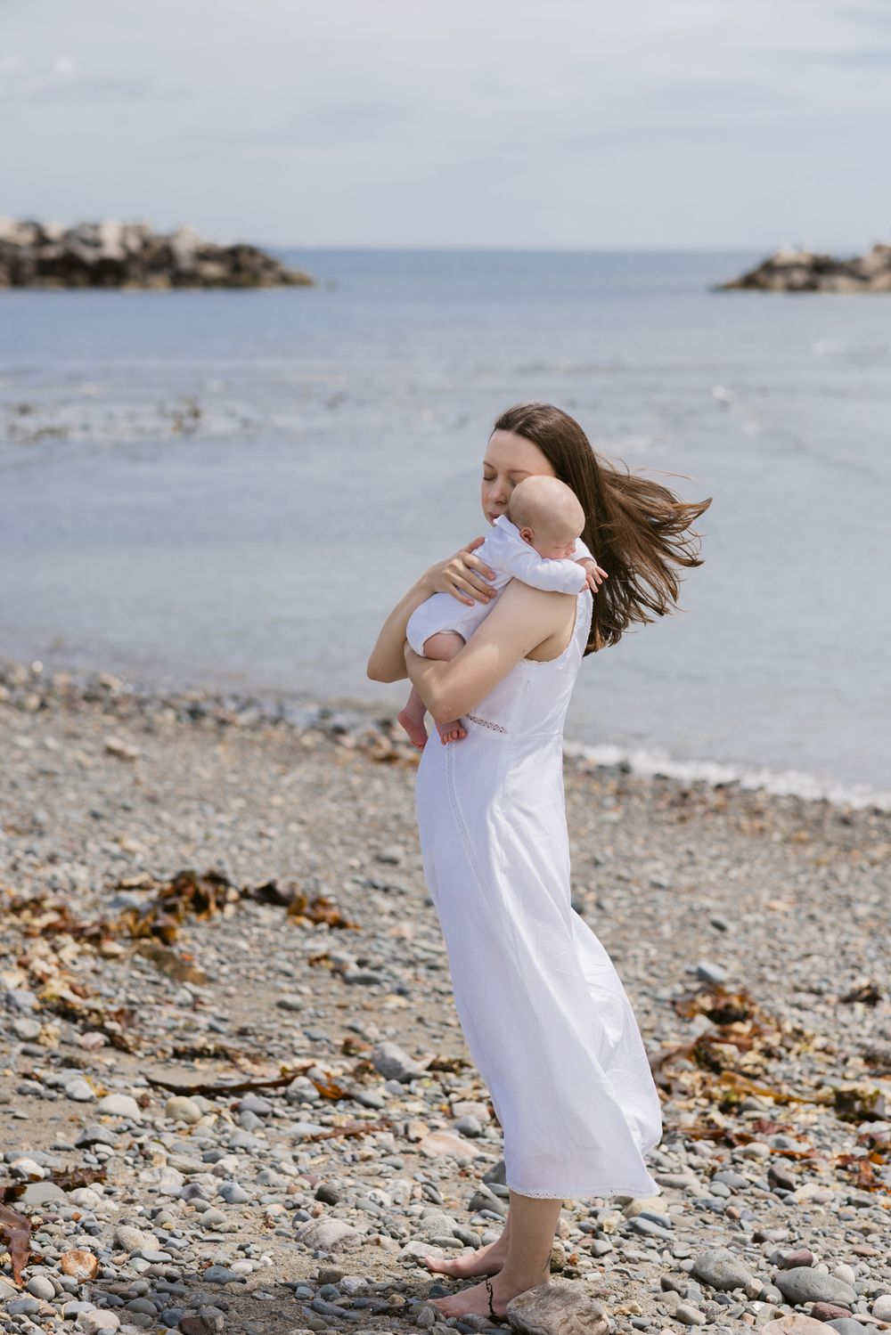 Mother wears white dress holding baby on rocky beach for newborn photos. Seacoast, Rye, Kennebunkport Hampton newborn photographer