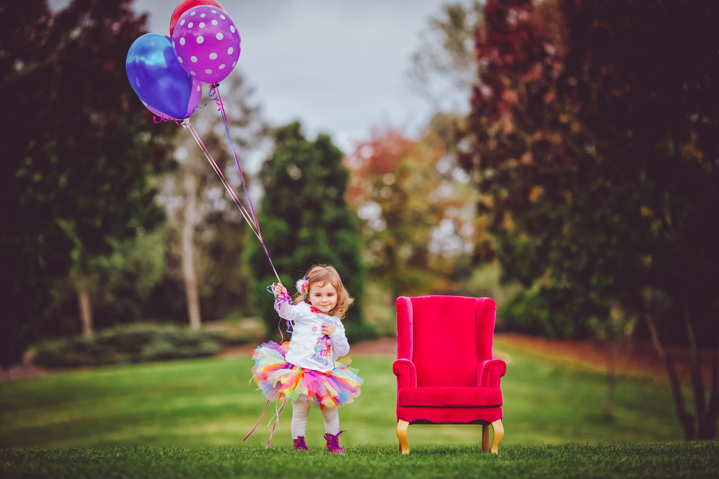 Toddler girl in tutu holding colorful balloons beside a small red armchair on green grass in a park.