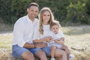 Happy family of three sitting outdoors on grass, parents in white shirts with toddler son, smiling in natural sunlight.