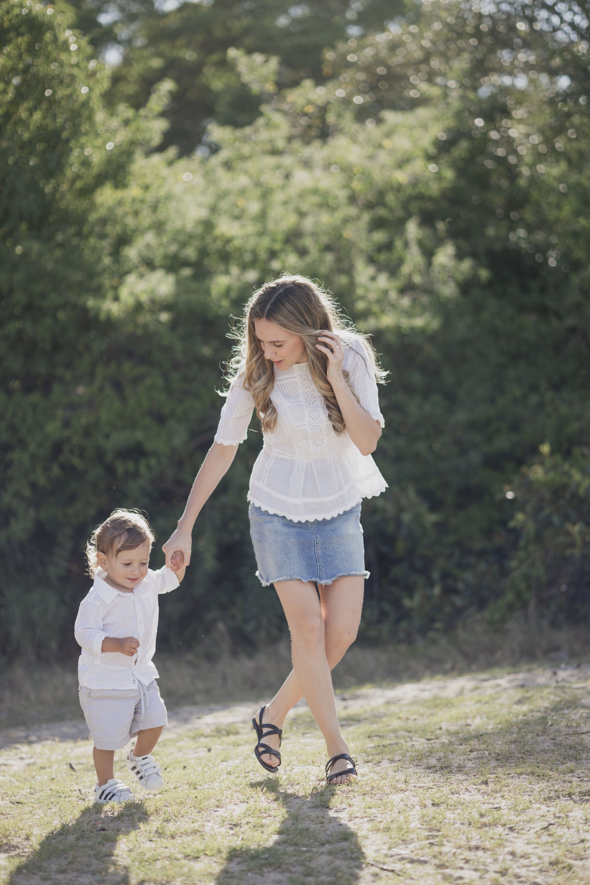A young mother in a white top and denim skirt holds hands with her toddler while walking outdoors in a sunny park.