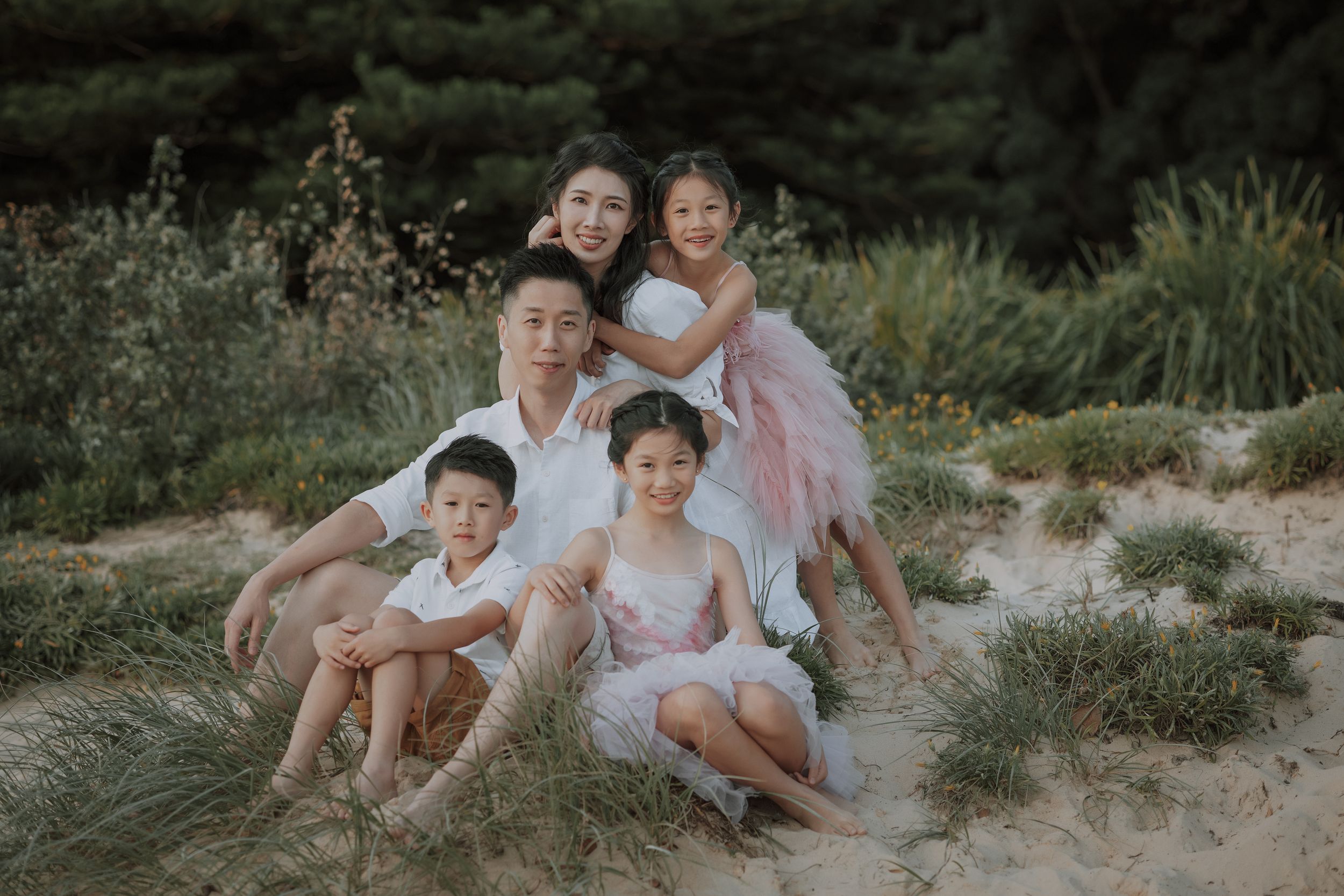 Asian family of five posing together on sandy beach dunes, parents with three children in white and pink outfits.