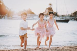 Three children run joyfully on a sandy beach at sunset, two girls in pink tutus and a boy in white shirt.