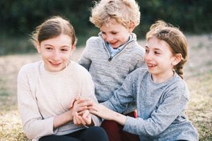 Three happy children sitting outdoors on grass, laughing and playing together on a sunny day.