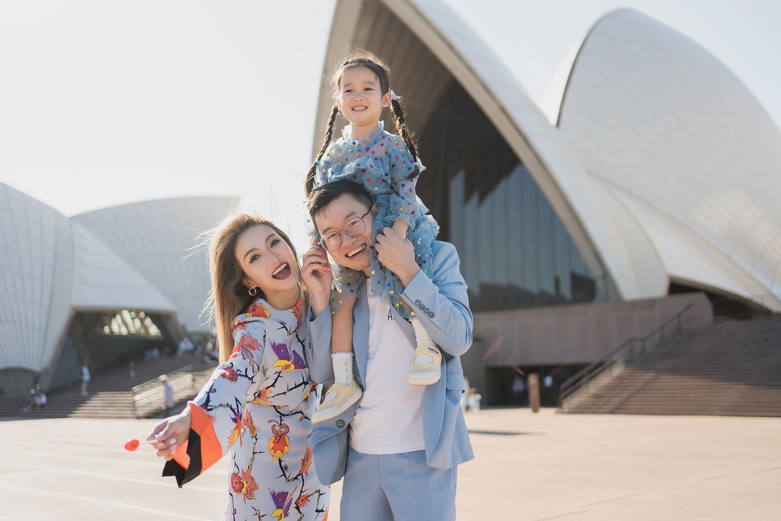 Family of three posing happily in front of Sydney Opera House on a sunny day.